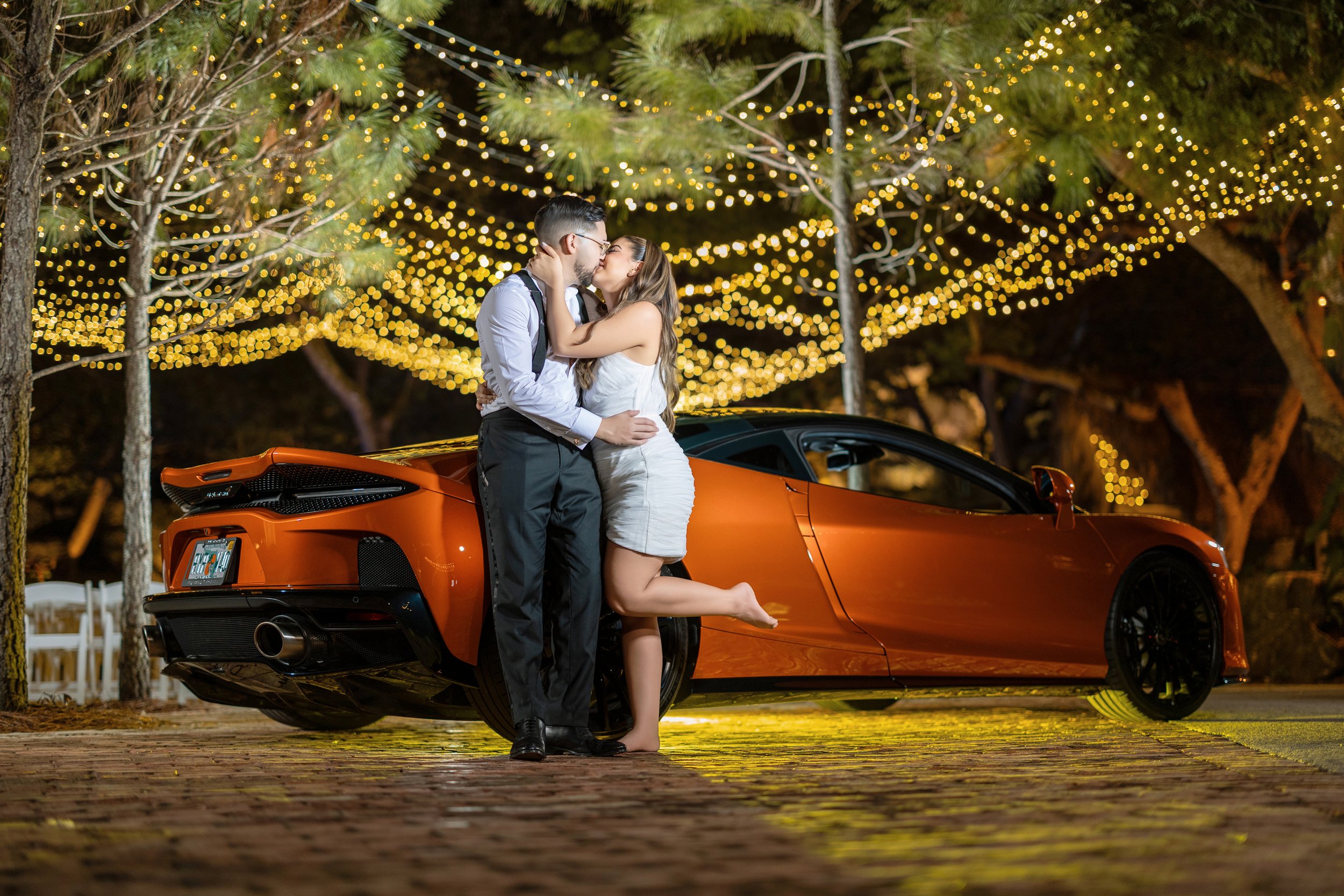 A couple kissing in front of a bright orange sports car under string lights hanging in trees during nighttime.