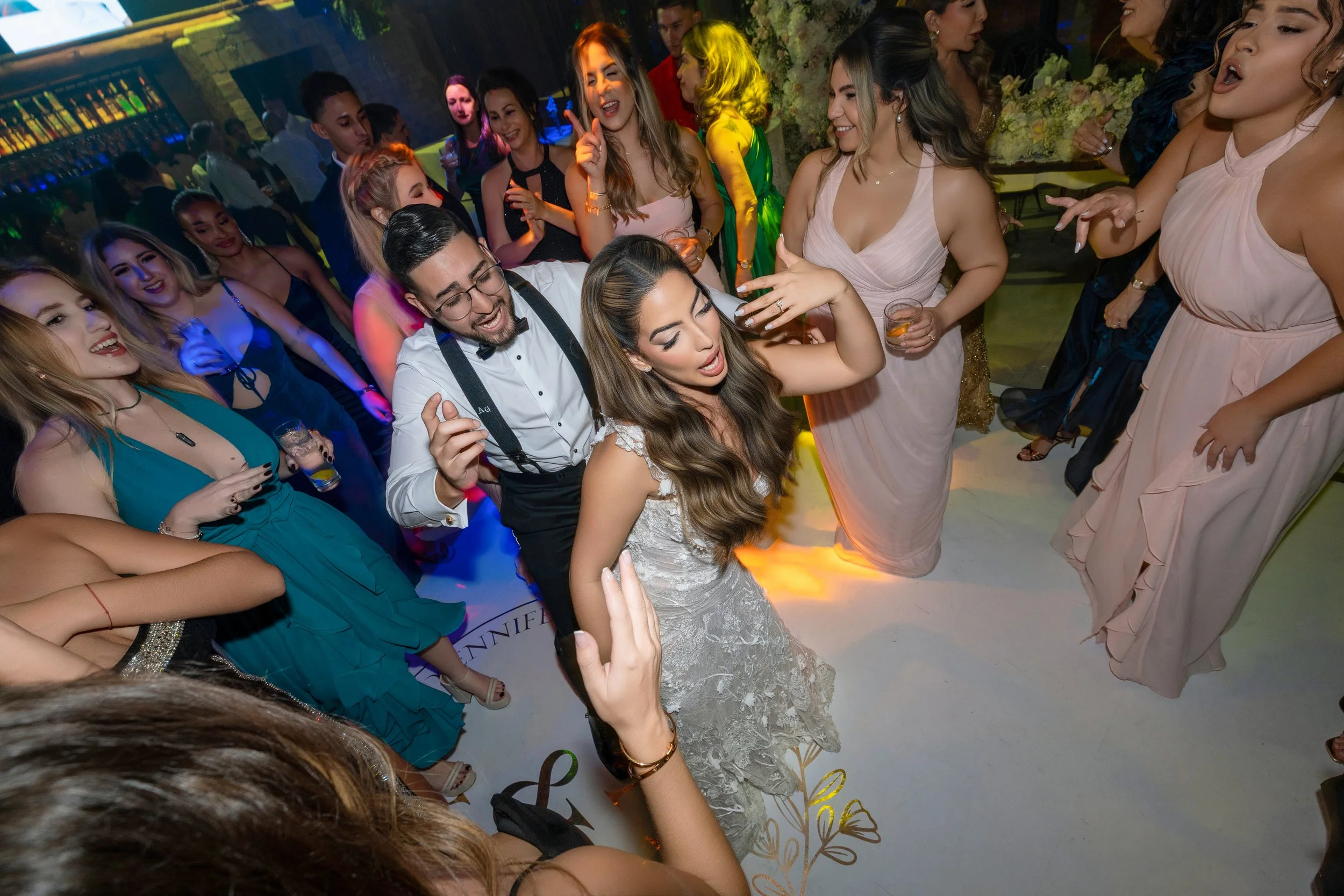 People dancing and having fun at a party or celebration, with a bride in a wedding dress and groom in formal attire in the center.