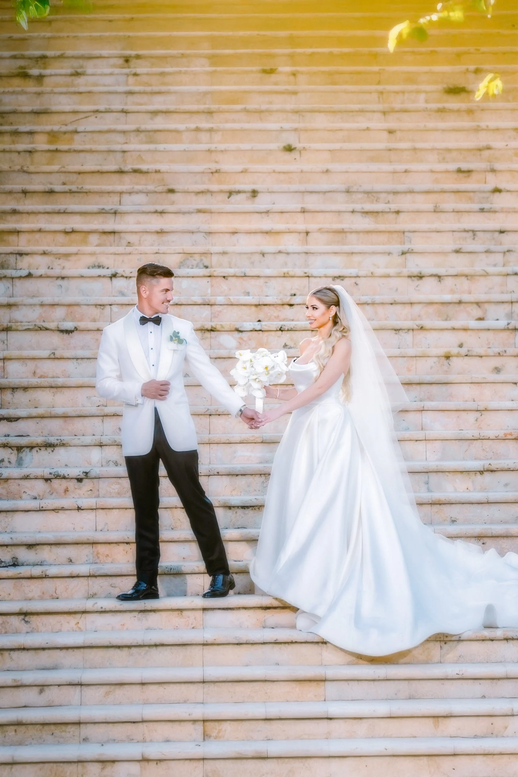 A bride and groom standing on a stone staircase, holding hands. The groom is in a white tuxedo jacket, black pants, and bow tie. The bride is in a white wedding gown with a long veil, holding a bouquet of white flowers.