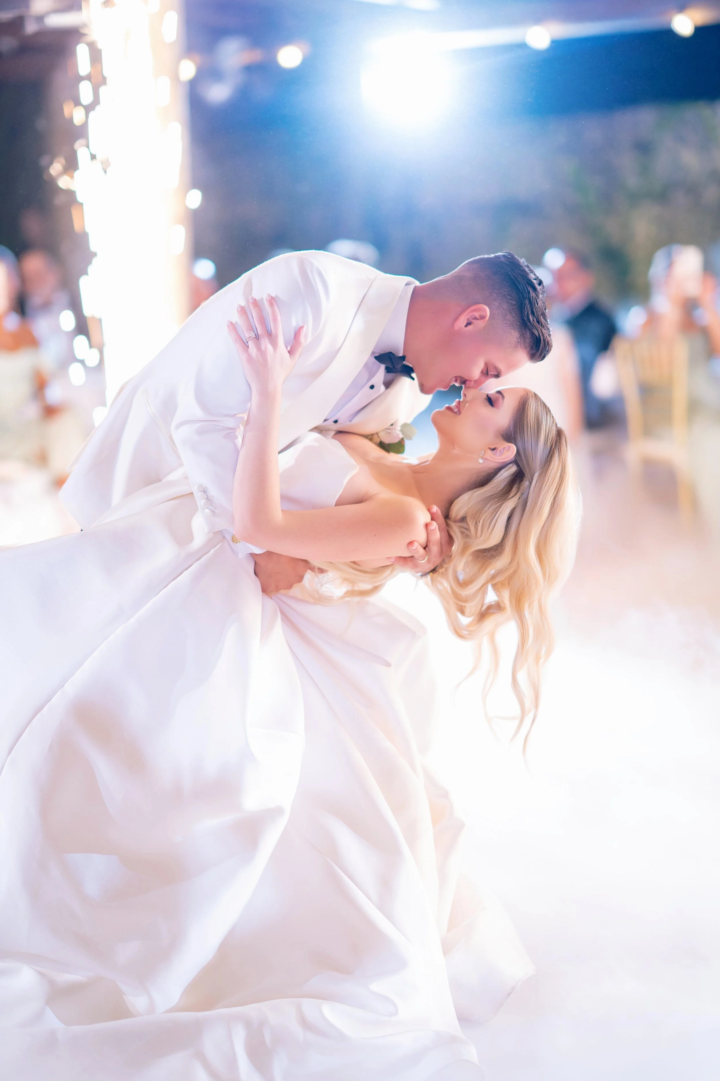 A bride and groom sharing a dance at their wedding reception, with the groom dipping the bride and both smiling. The background shows blurred guests and sparkling decorations.