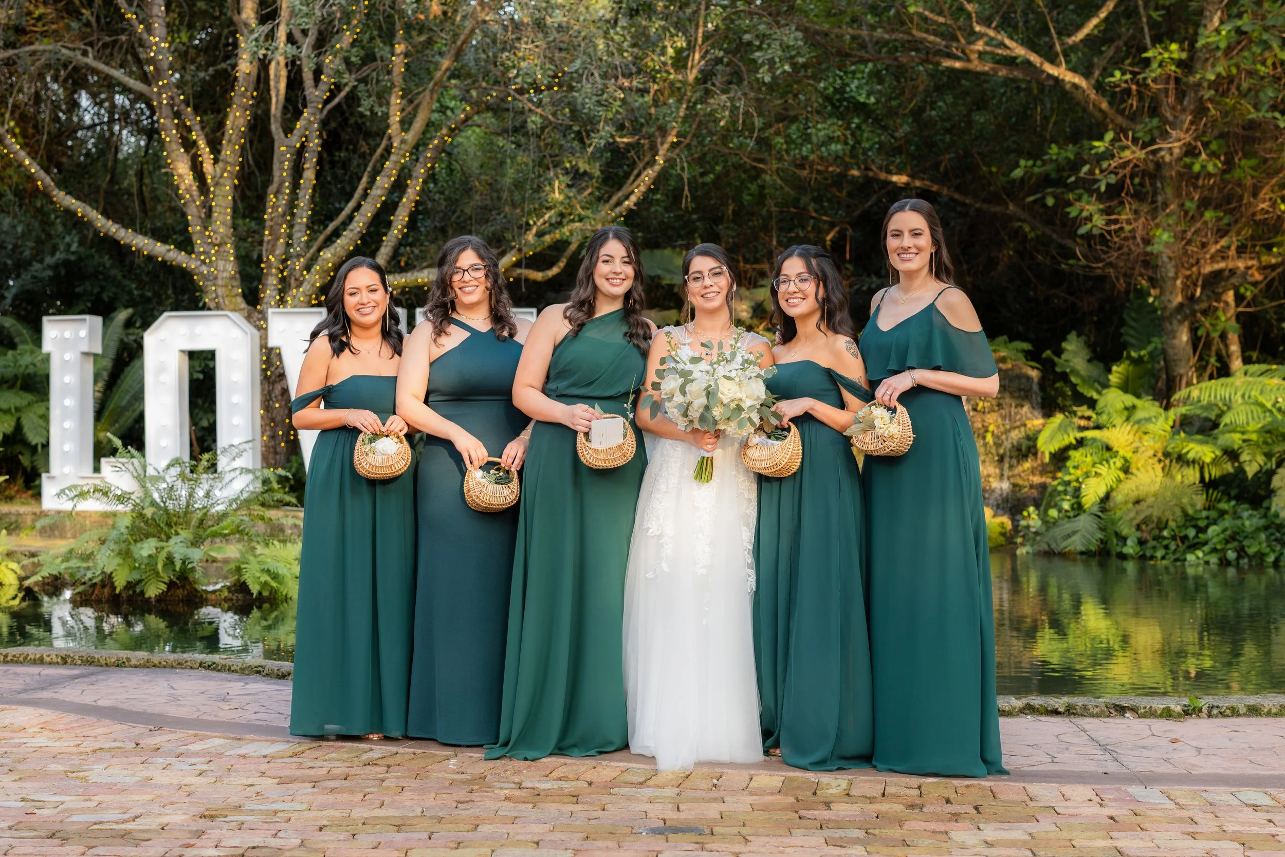 Group of six women in green dresses at a wedding celebration, holding baskets, with a bride in a white gown holding a bouquet of white flowers, standing outdoors amidst trees, decorative lights, and large illuminated letters '10'.