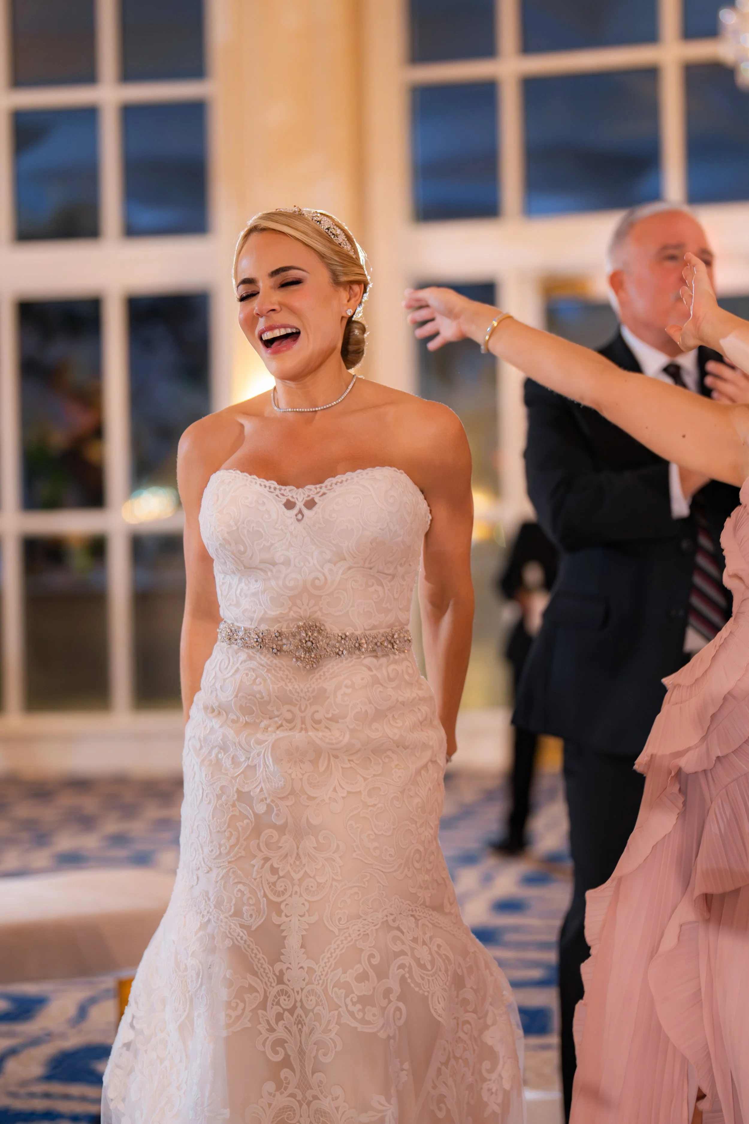 A bride in a strapless white wedding dress, smiling and laughing at her wedding reception, surrounded by guests.
