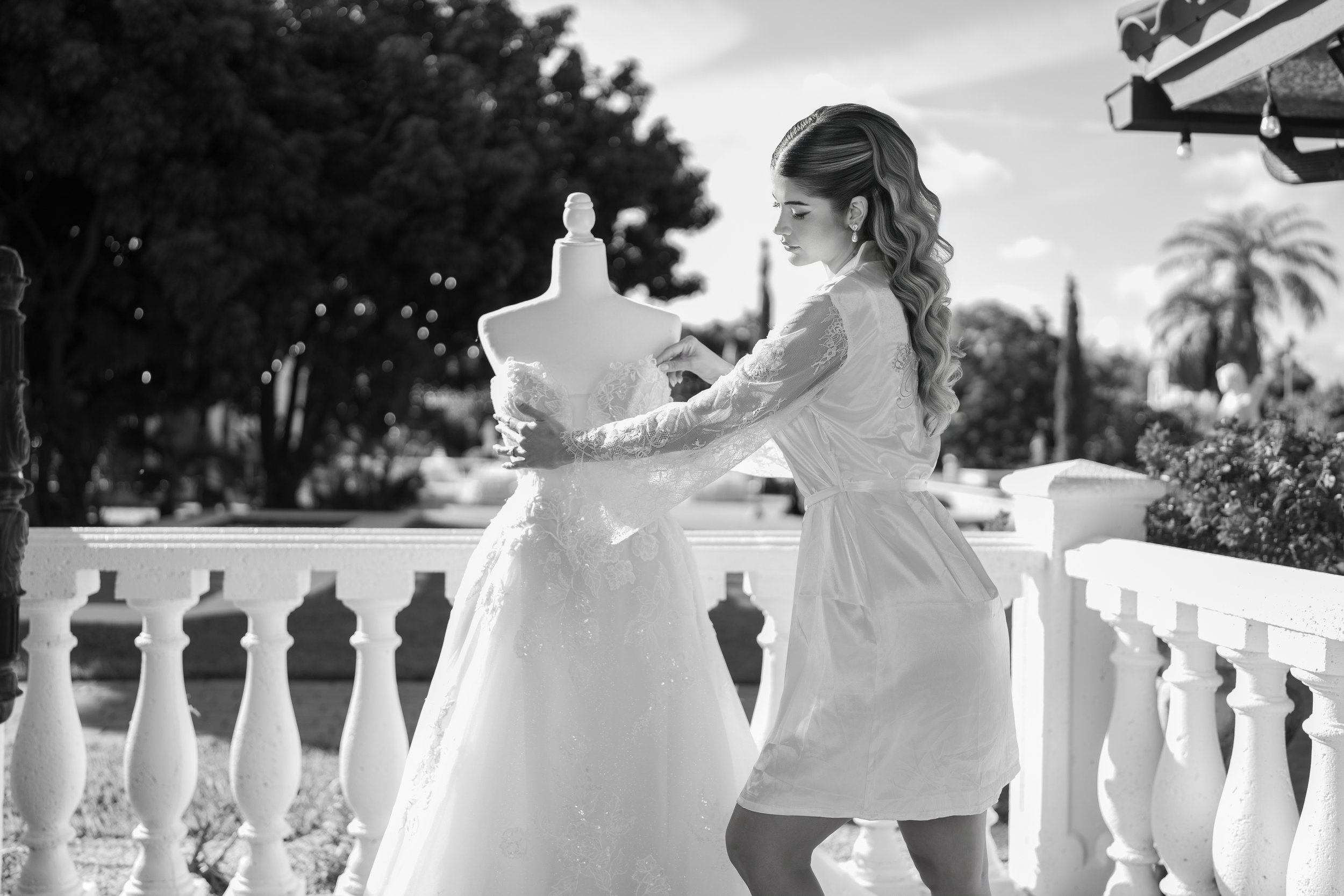 A woman in a satin robe is looking at a wedding dress on a mannequin outside on a balcony, with trees and palm trees in the background.