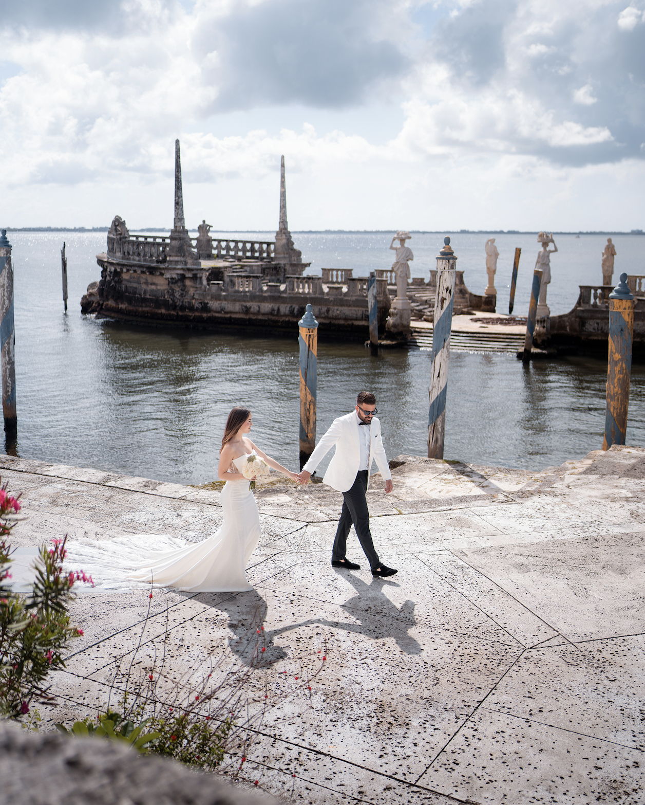A bride and groom walking hand-in-hand along a waterfront promenade during their wedding photoshoot, with an old, abandoned shipwreck and cloudy sky in the background.