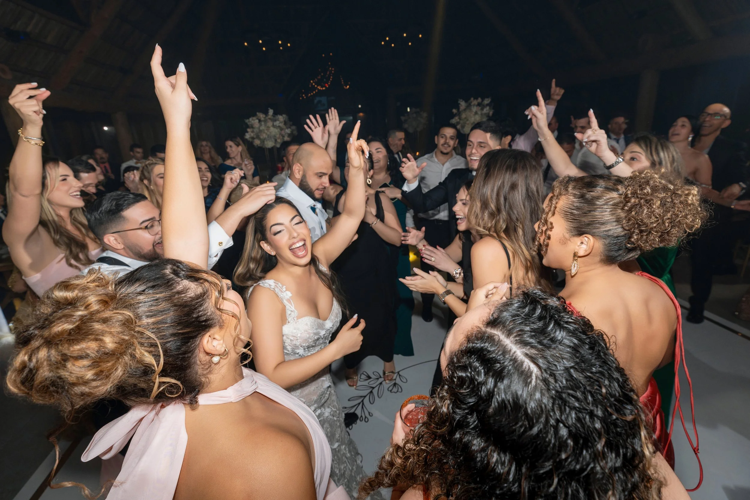 People dancing and celebrating at a wedding reception in a rustic venue with string lights and floral centerpieces.