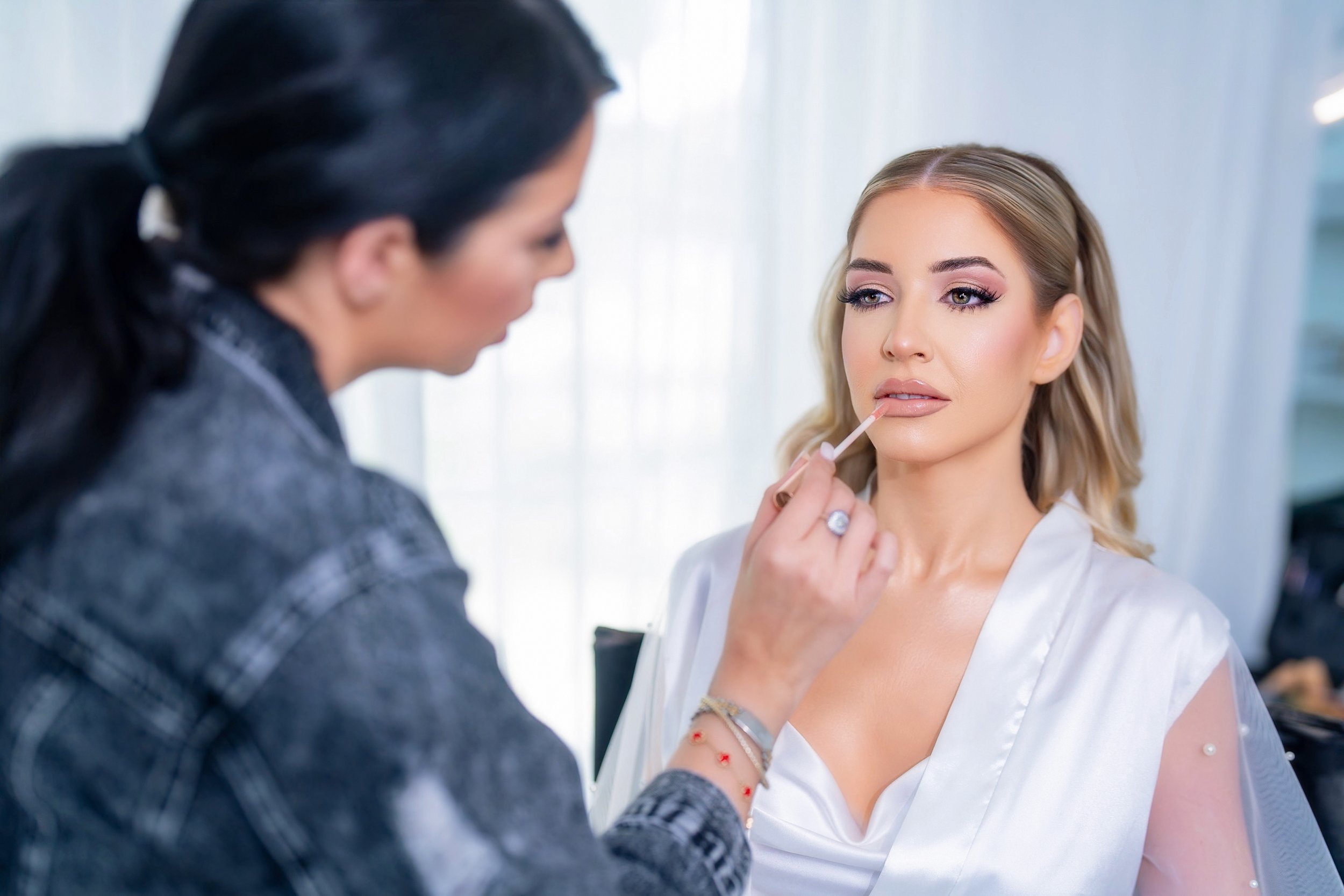 Makeup artist applying lip gloss on a woman with styled hair and makeup, sitting in a well-lit room.