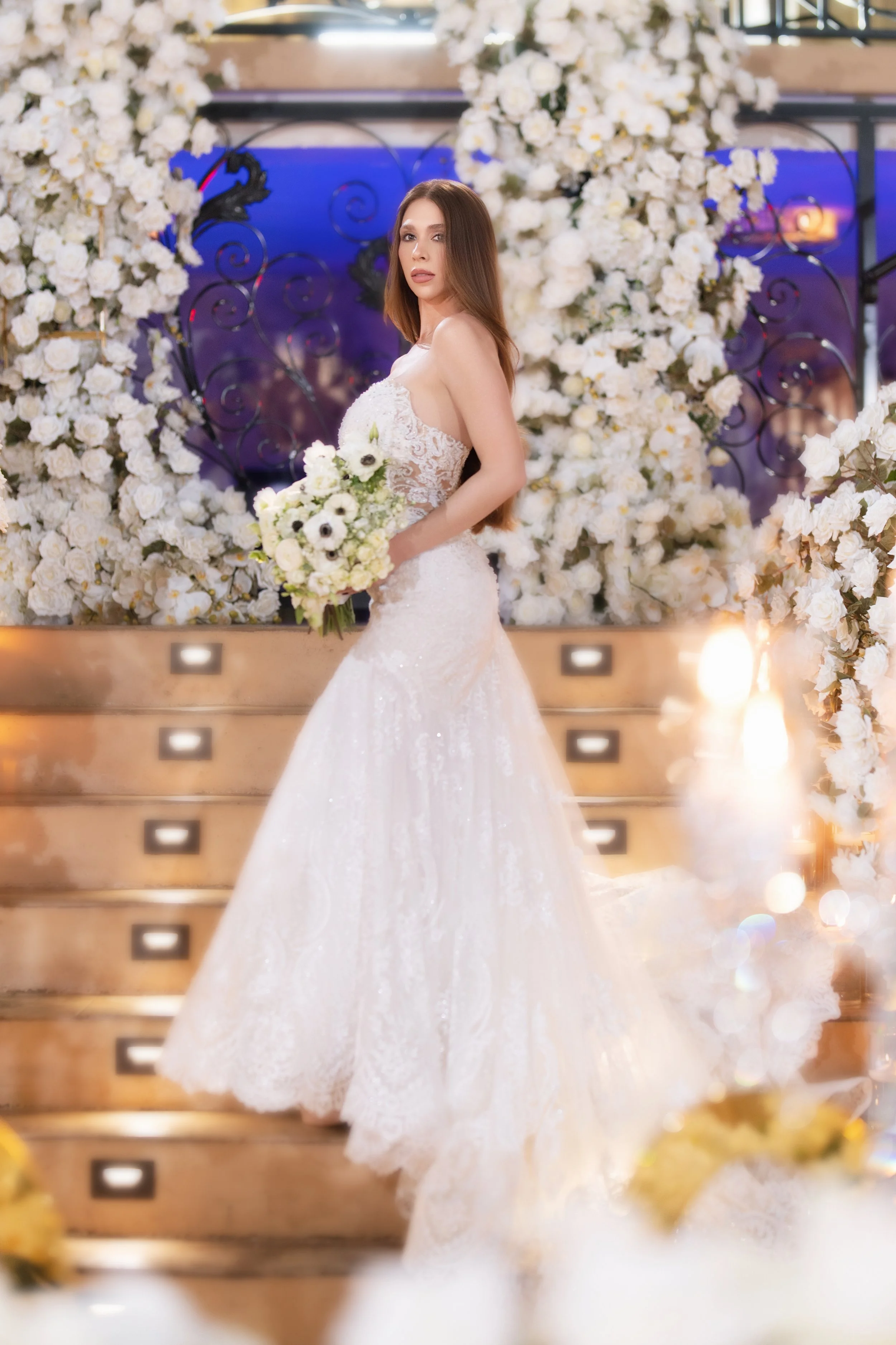 A woman in a white wedding dress holding a bouquet of white flowers, standing on a staircase decorated with white flowers and surrounded by floral arrangements at a wedding or event venue.