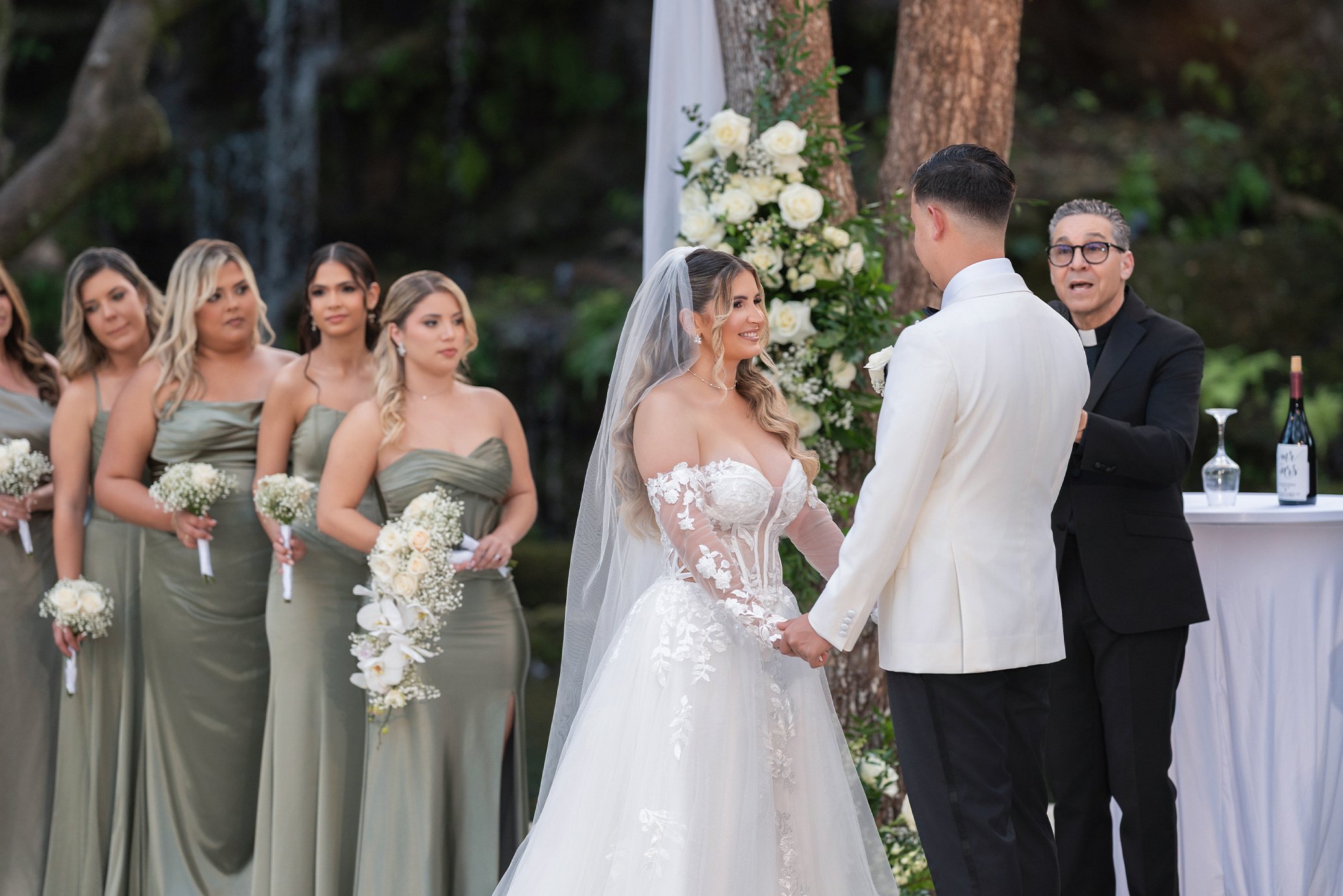 A wedding ceremony taking place outdoors, with a bride and groom holding hands and smiling at each other, surrounded by bridesmaids in green dresses, an officiant, and a decorated backdrop with flowers and trees.