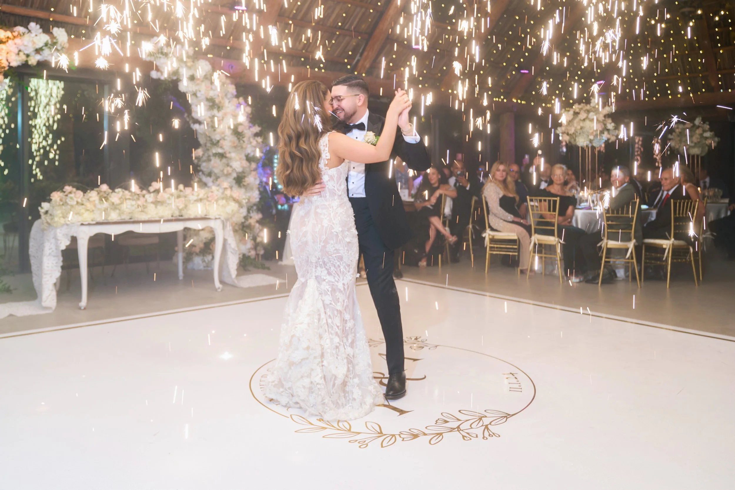 A bride and groom sharing their first dance at their wedding reception, surrounded by sparklers with guests seated at tables in the background.