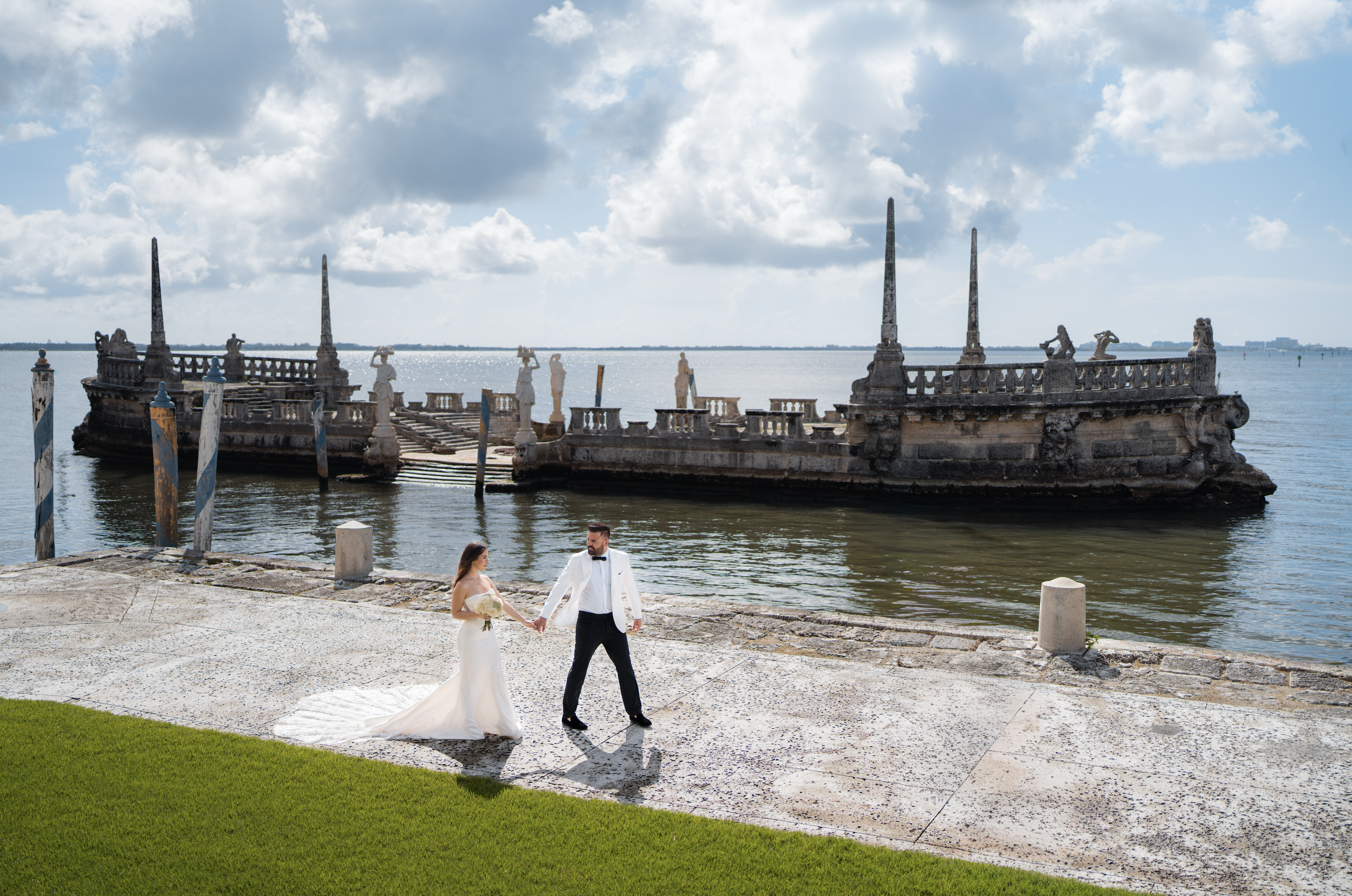 A bride and groom holding hands by a lakeside with an ornate, historic stone structure with sculptures and spires in the background.