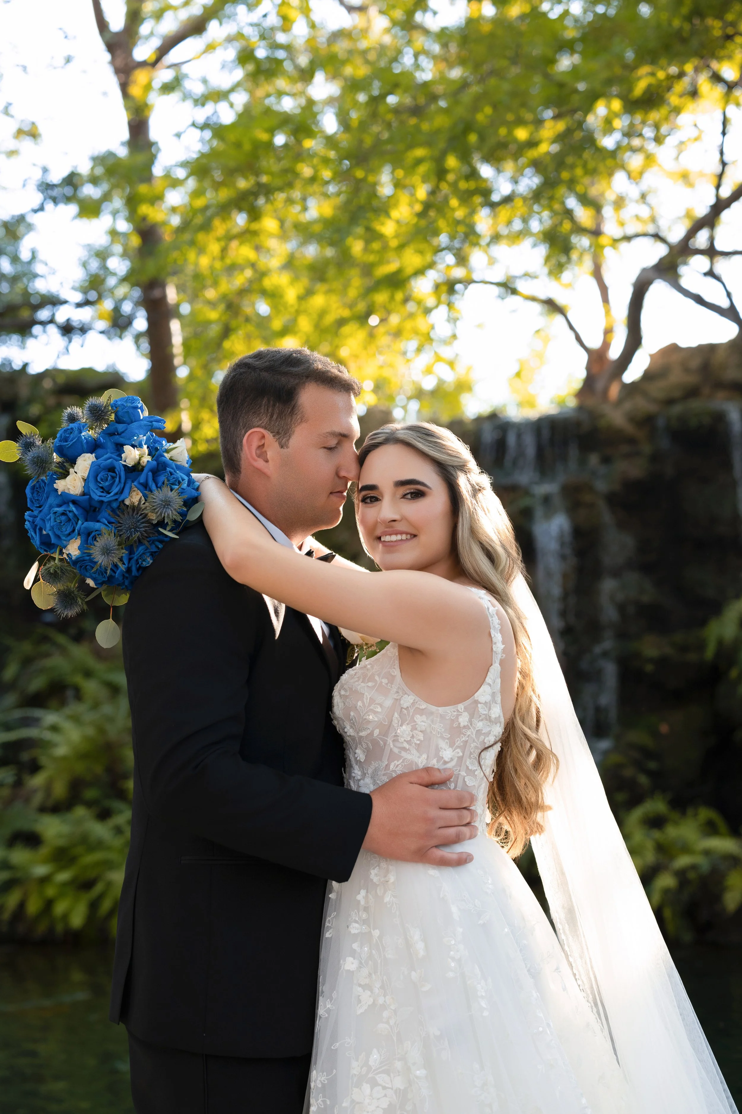 A bride and groom embrace outdoors with a waterfall and lush trees in the background, daylight shining through.