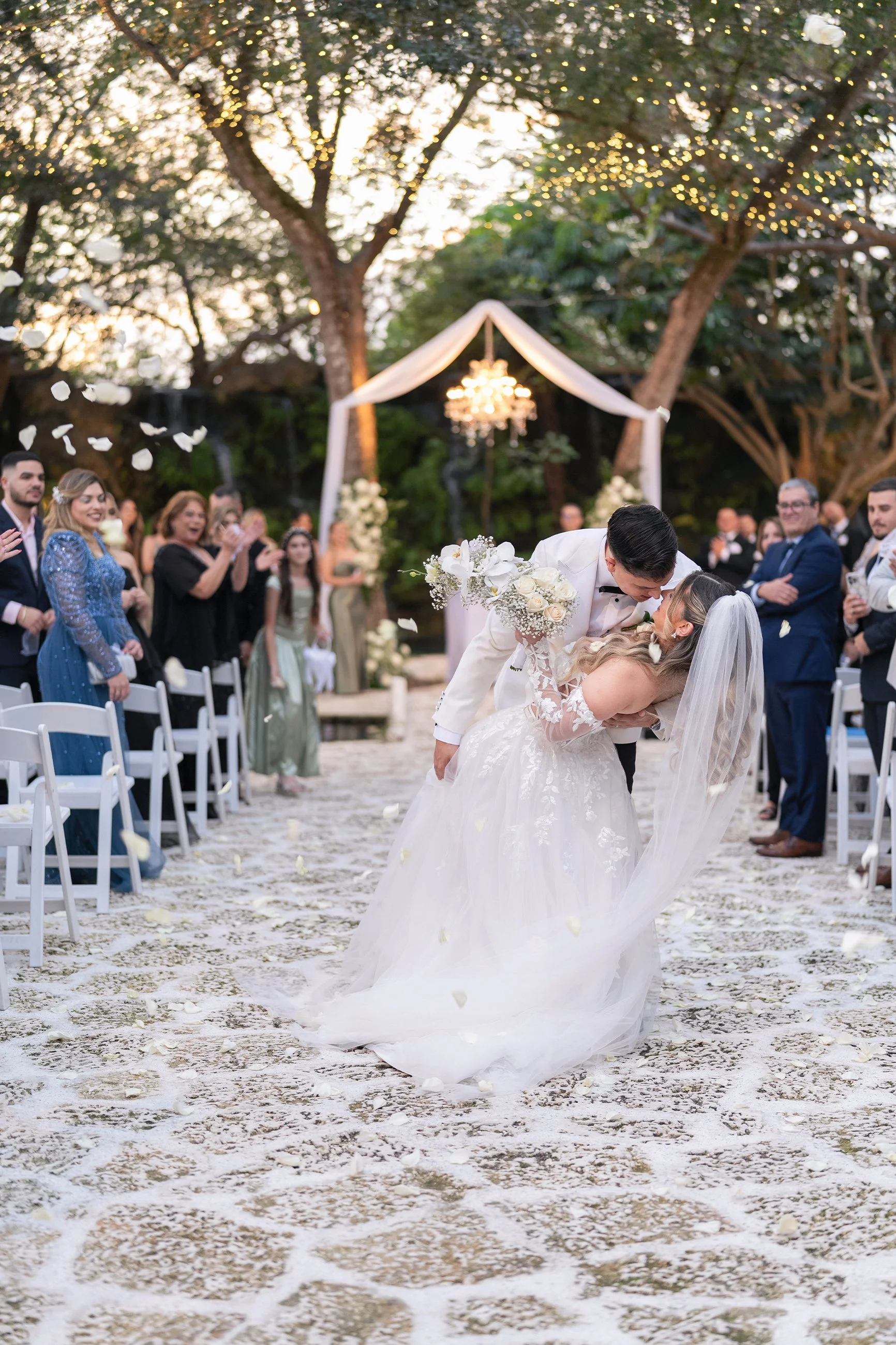 A newlywed couple shares a kiss during their outdoor wedding ceremony as guests applaud. The bride is in a white wedding gown with a veil, holding a bouquet, and the groom is in a tuxedo. Heart-shaped petals are falling around them, and there is a de