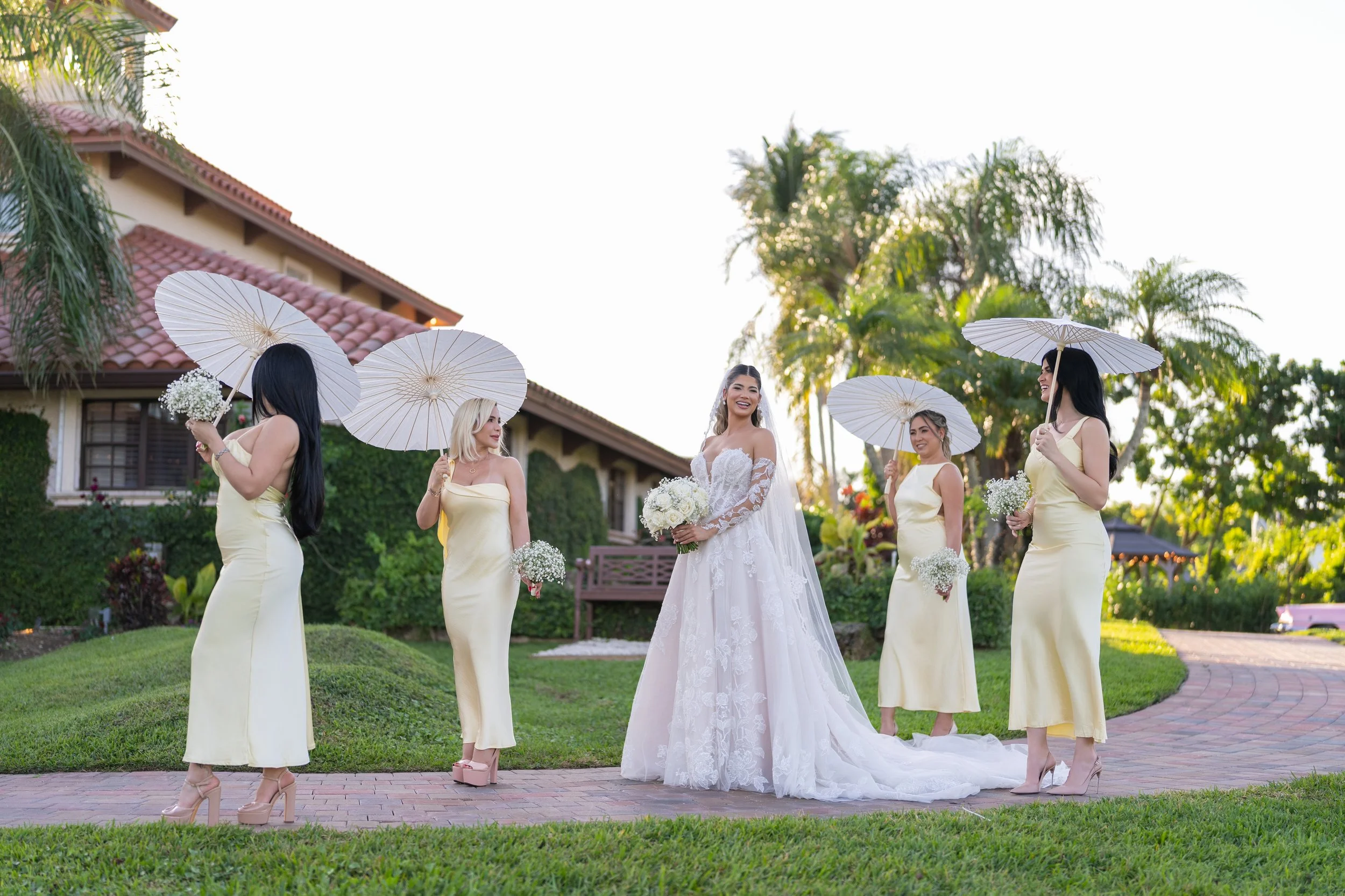 A bride in a white wedding gown holding a bouquet of white flowers stands outdoors on a brick pathway, surrounded by four bridesmaids in yellow dresses holding white parasols and small bouquets, in a garden with trees and a house in the background.