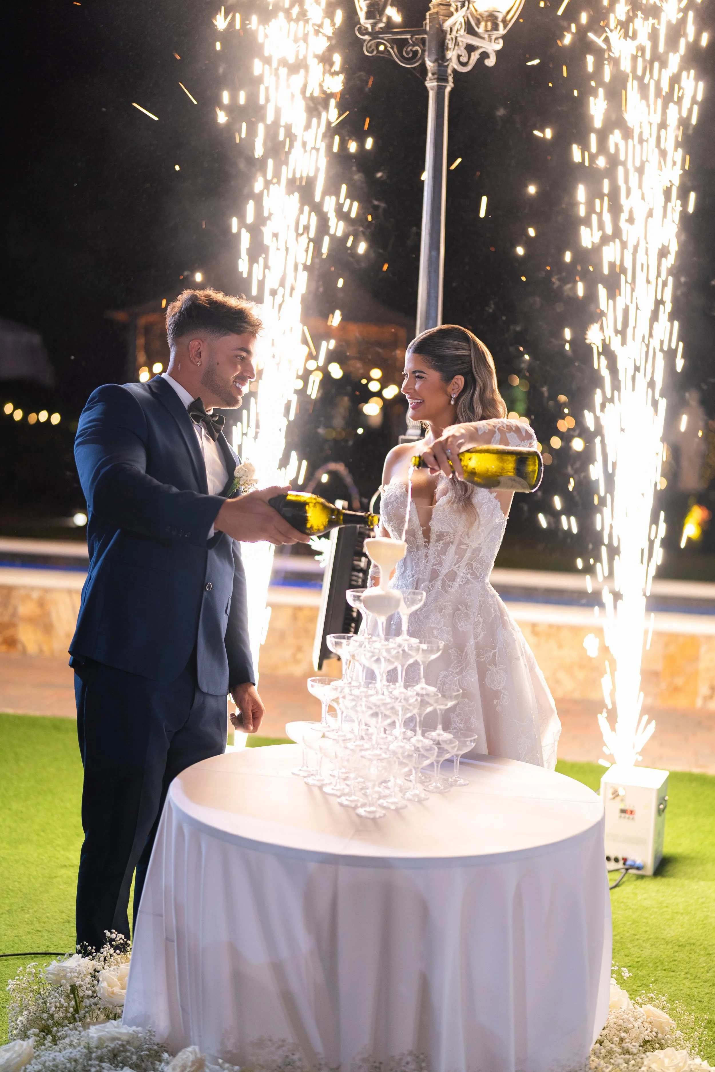 A newlywed couple in wedding attire celebrating at night with a champagne tower and fireworks.