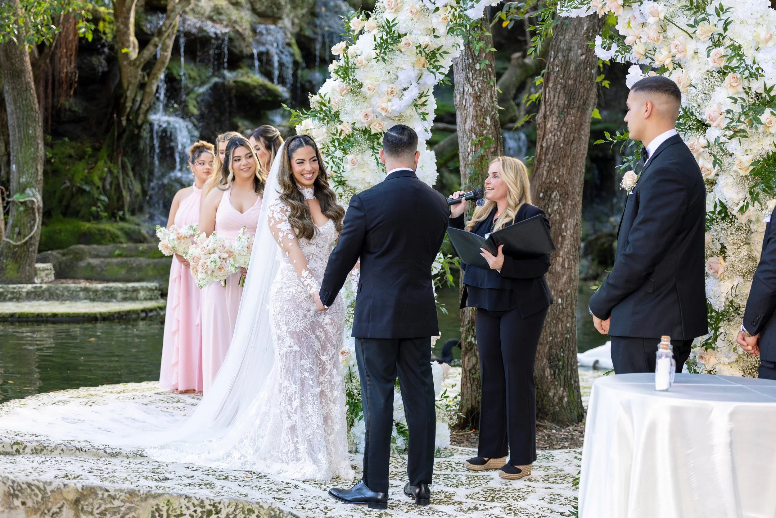 A wedding ceremony taking place outdoors by a waterfall, with a bride and groom holding hands, standing in front of an officiant. Bridesmaids in pink dresses and groomsmen in black suits are standing nearby, with a floral arch decorated with white an
