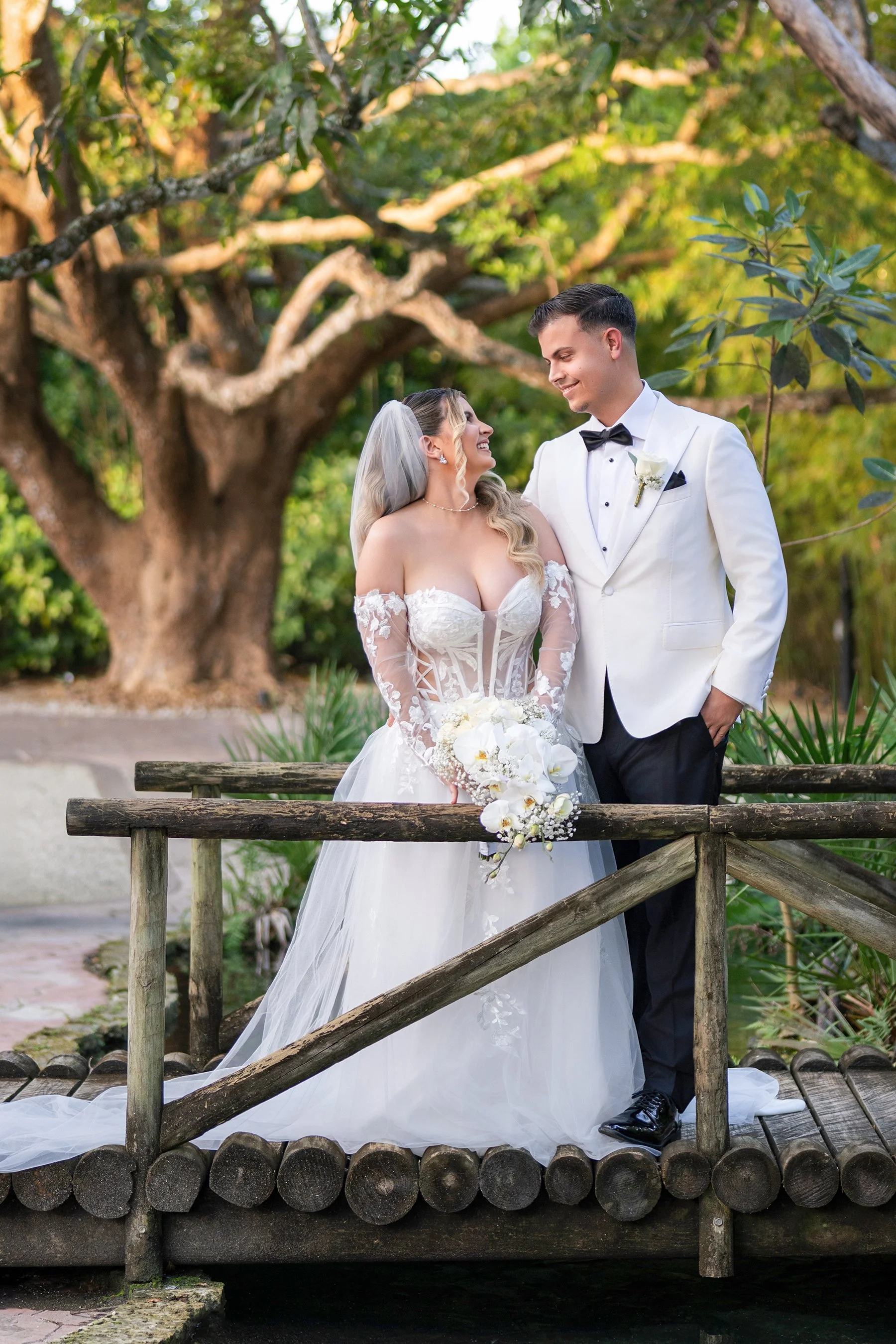 Bride and groom standing on a small wooden bridge outdoors, gazing at each other, surrounded by greenery and large trees, with the bride holding a white floral bouquet.
