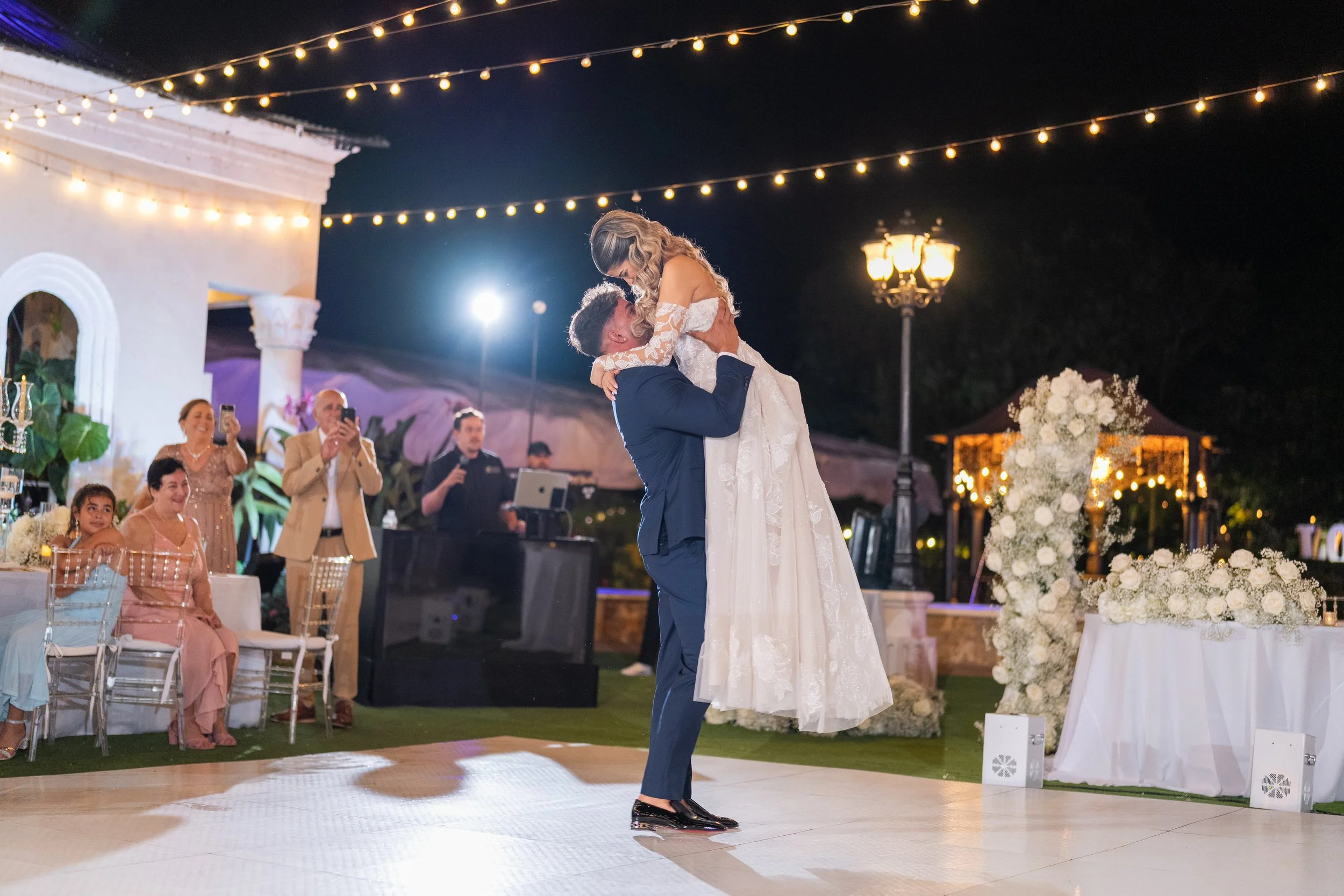 A bride and groom dancing at their wedding reception outside at night, with guests watching and taking photos, decorated with string lights and flower arrangements.