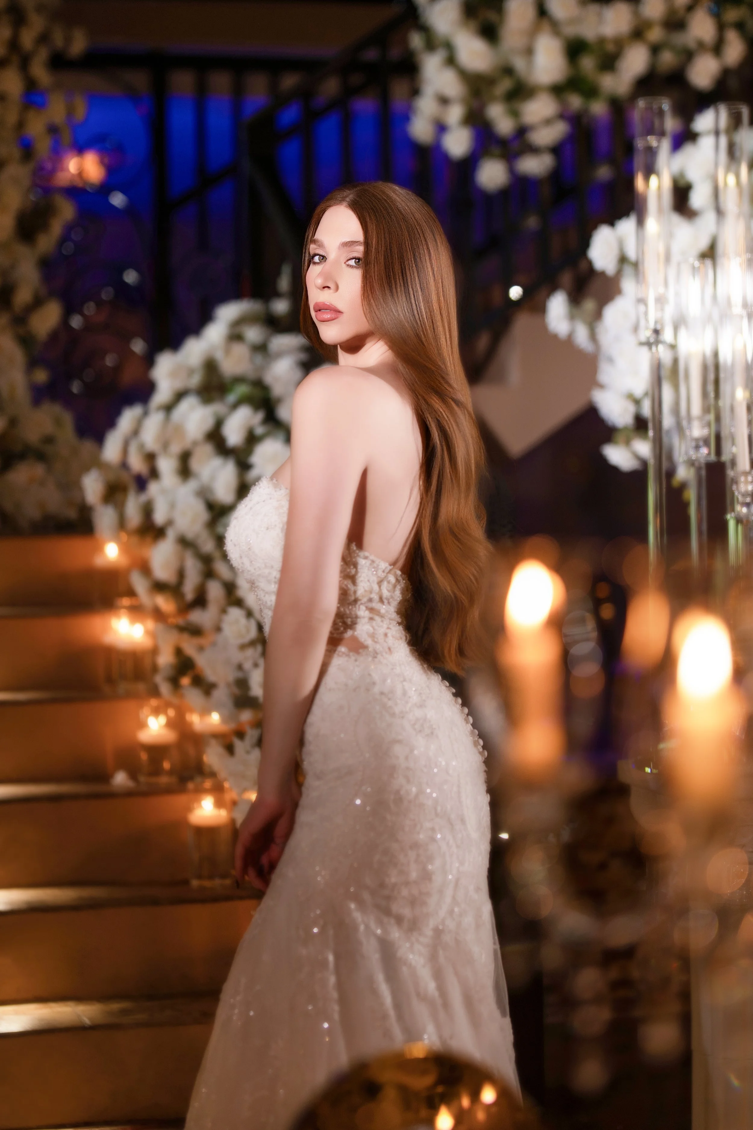 A woman in a wedding dress standing indoors near candles and floral decorations, with a staircase in the background.