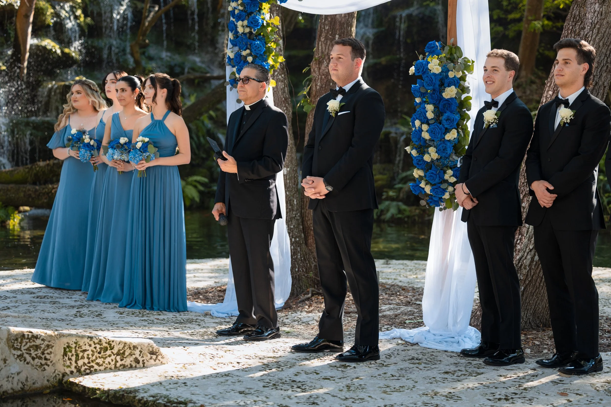 A wedding ceremony outdoors by a waterfall with six people standing in a row. Three women in blue dresses, two men in black tuxedos, and an officiant in black with glasses. The scene is decorated with blue and white flowers on a white fabric backdrop