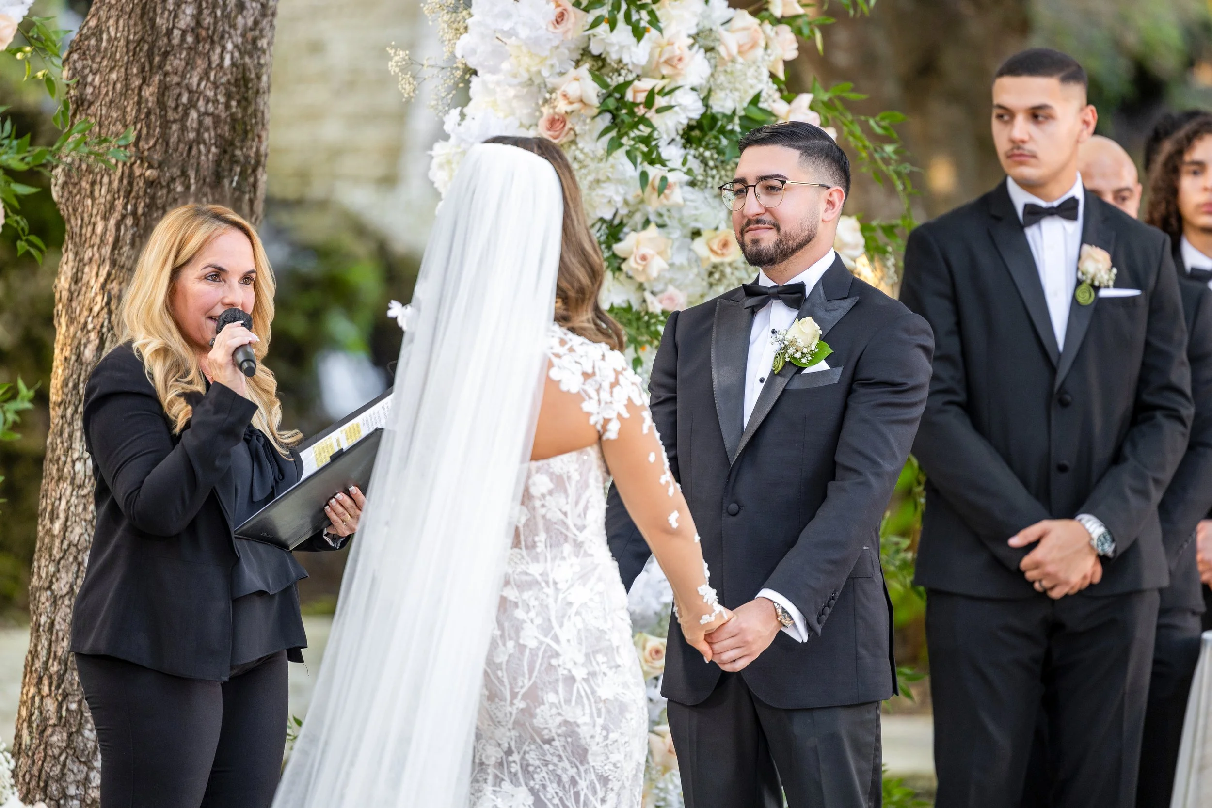 A couple getting married outdoors, holding hands and facing each other, with an officiant woman speaking into a microphone. The bride wears a white lace gown and veil, and the groom wears a black tuxedo with a bow tie and boutonniere. There is a flor