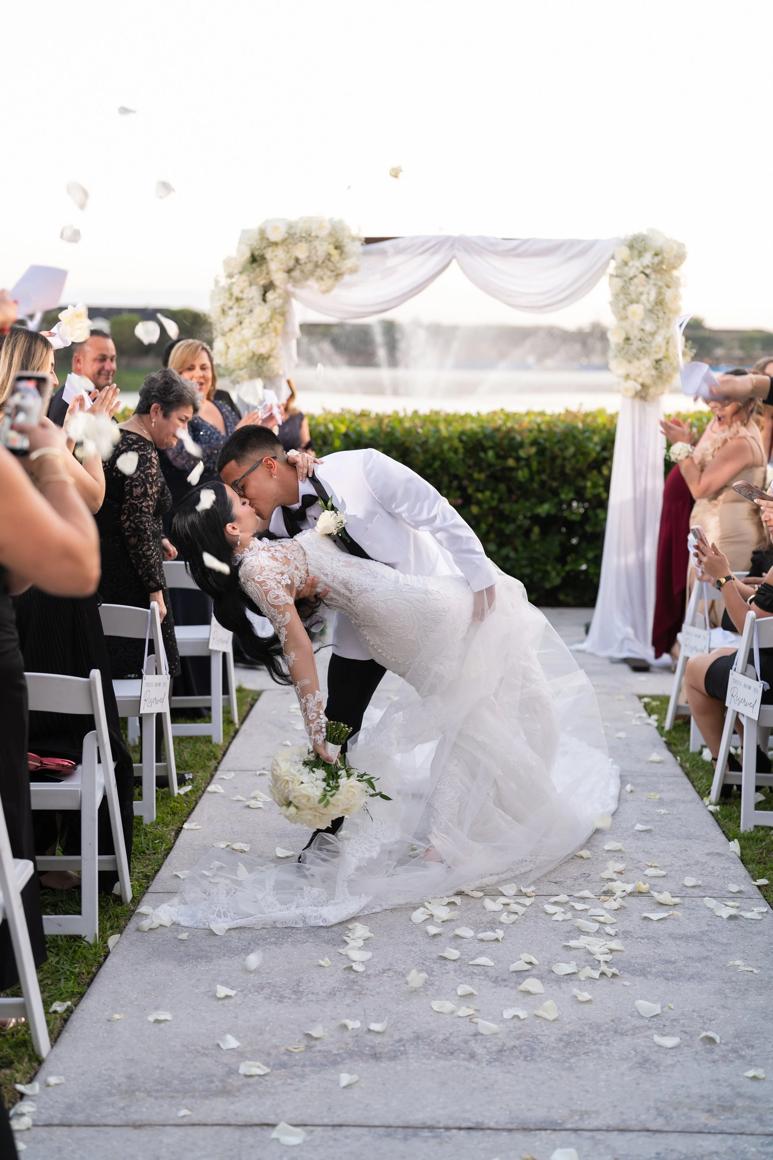 A newlywed couple sharing a kiss during their outdoor wedding ceremony, with guests applauding and throwing white flower petals, and a decorative floral arch in the background.