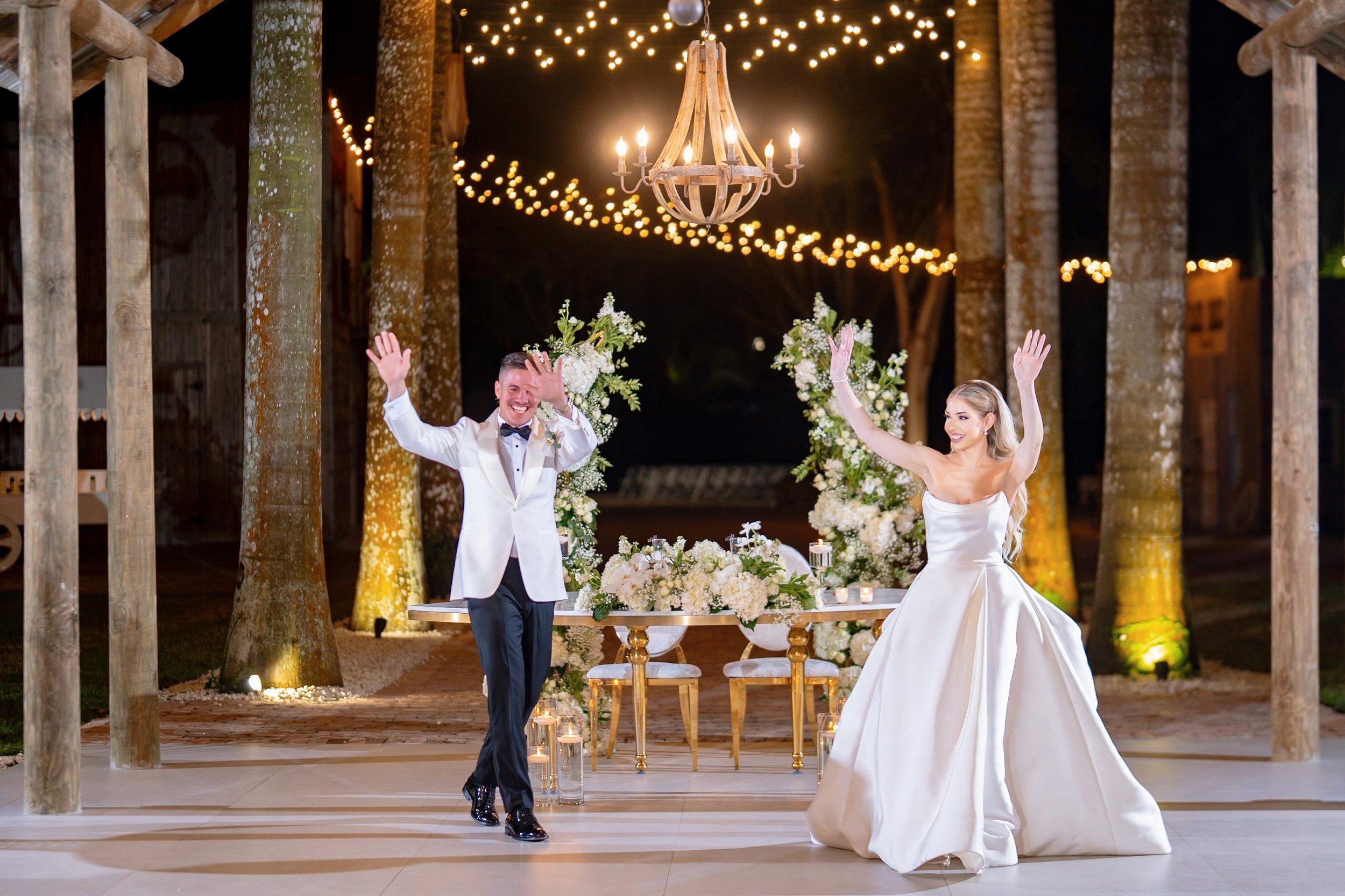 Bride and groom dancing at their wedding reception, dressed in formal attire, under hanging lights, with floral decorations in the background.