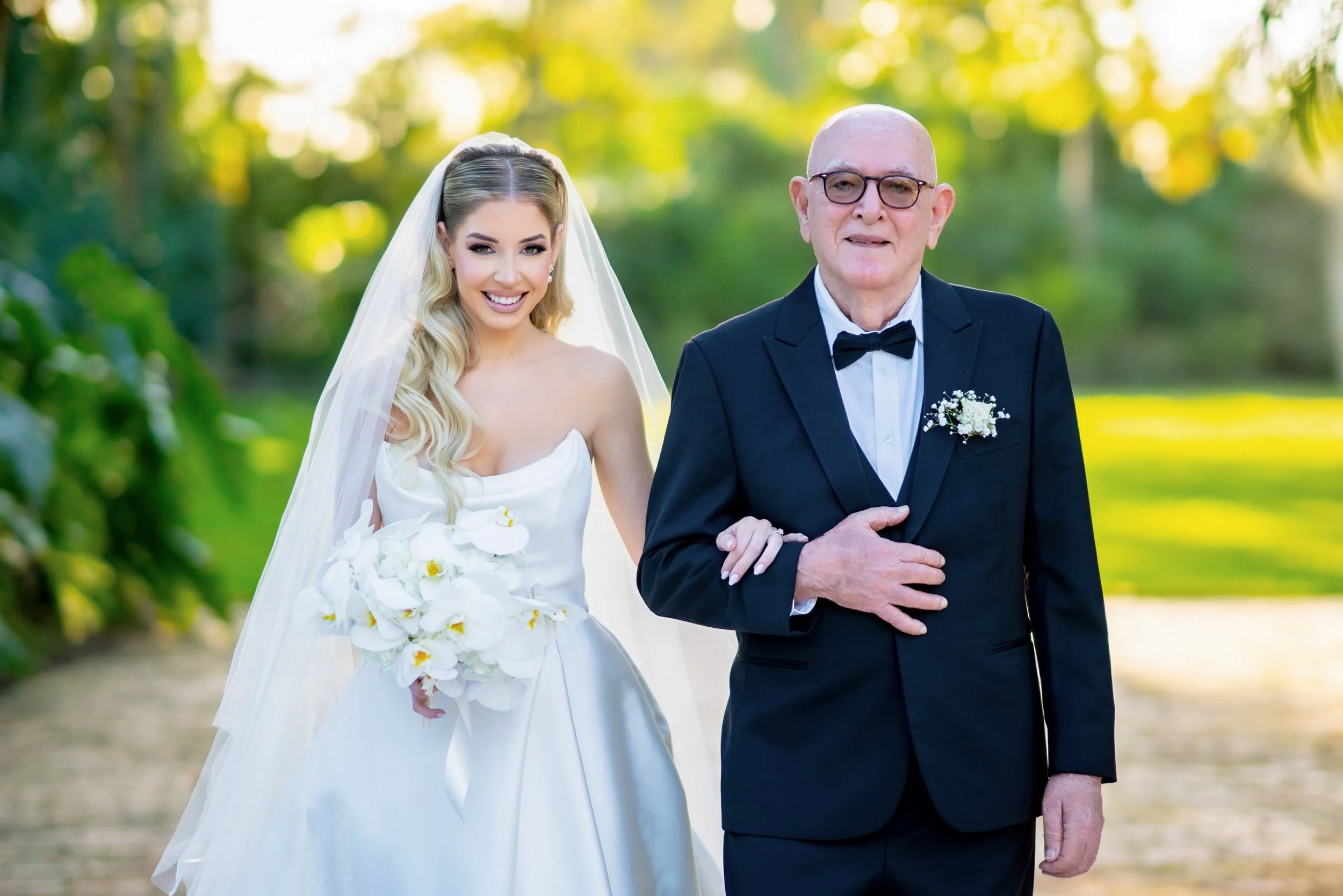 A bride in a white wedding gown and veil holding a bouquet of white orchids walks arm-in-arm with a man in a black tuxedo and bow tie, outdoors with green trees and sunlight in the background.