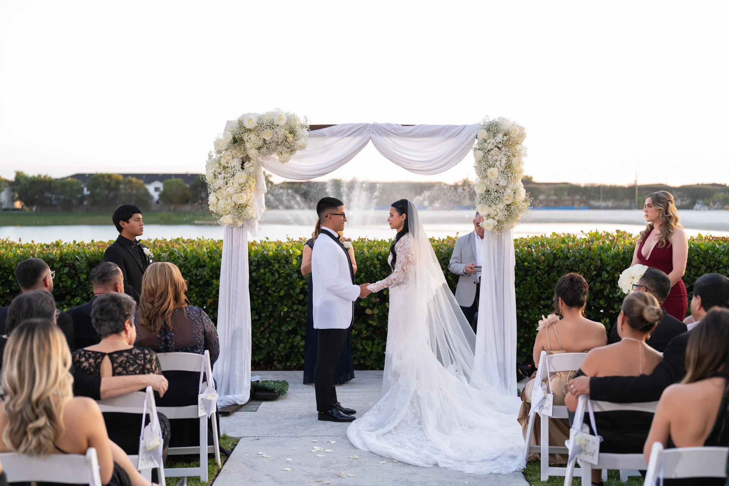 A wedding ceremony outdoors by a lake with the bride and groom holding hands, under a white floral arch, surrounded by seated guests.