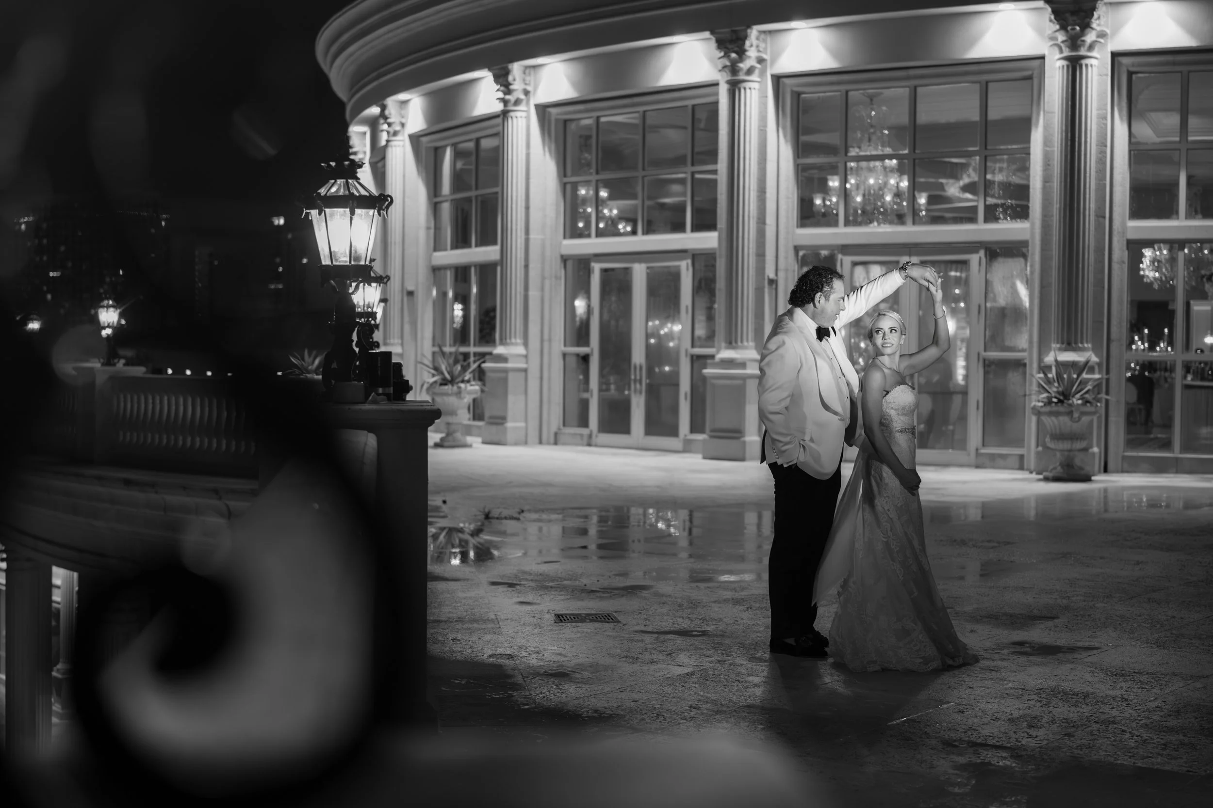 A man and woman dancing romantically outside a grand building at night, illuminated by street lamps and chandeliers visible through the windows.