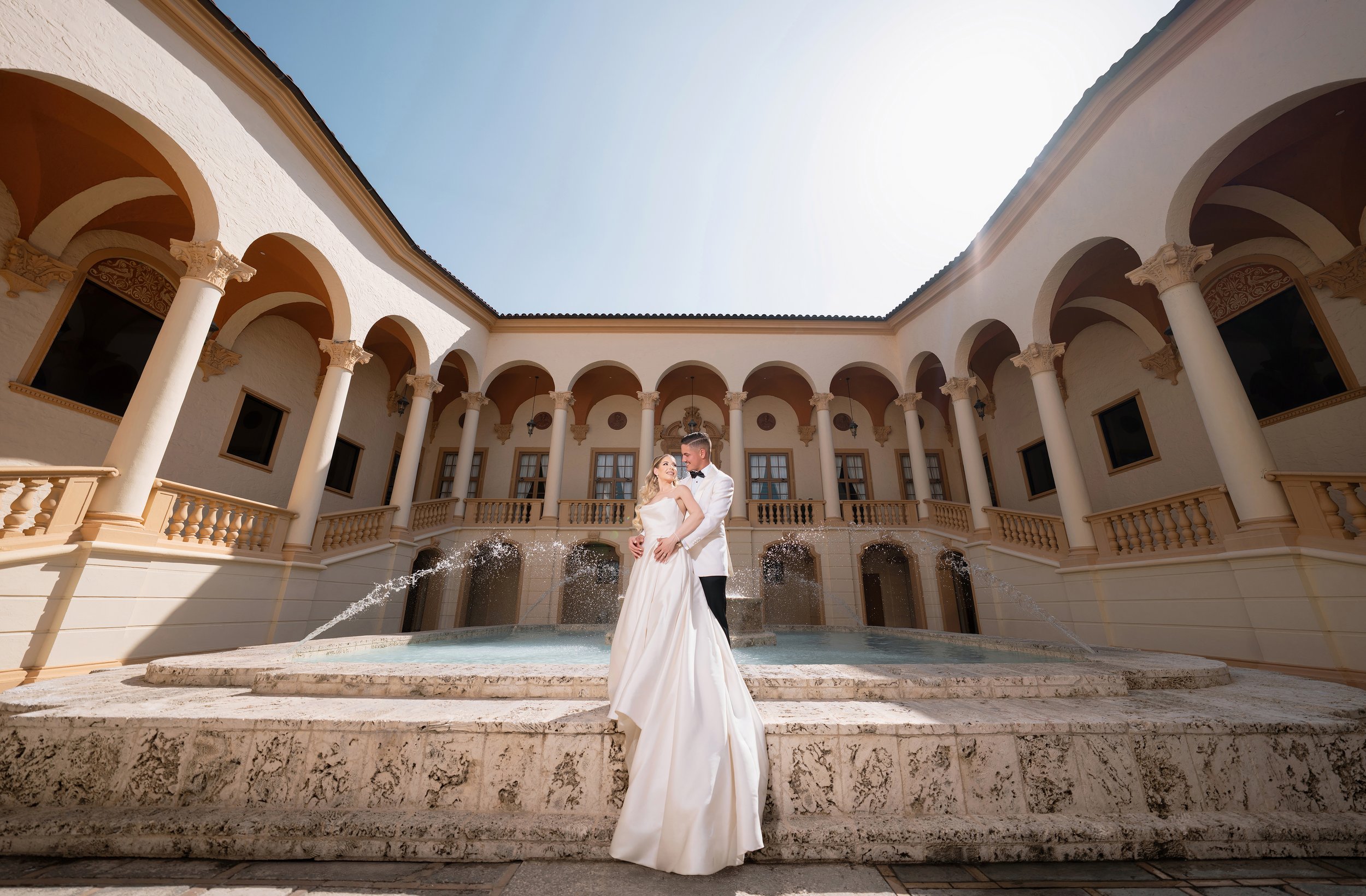 A couple in wedding attire standing in a fountain in a courtyard with classical architecture, columns, and a clear sky above.