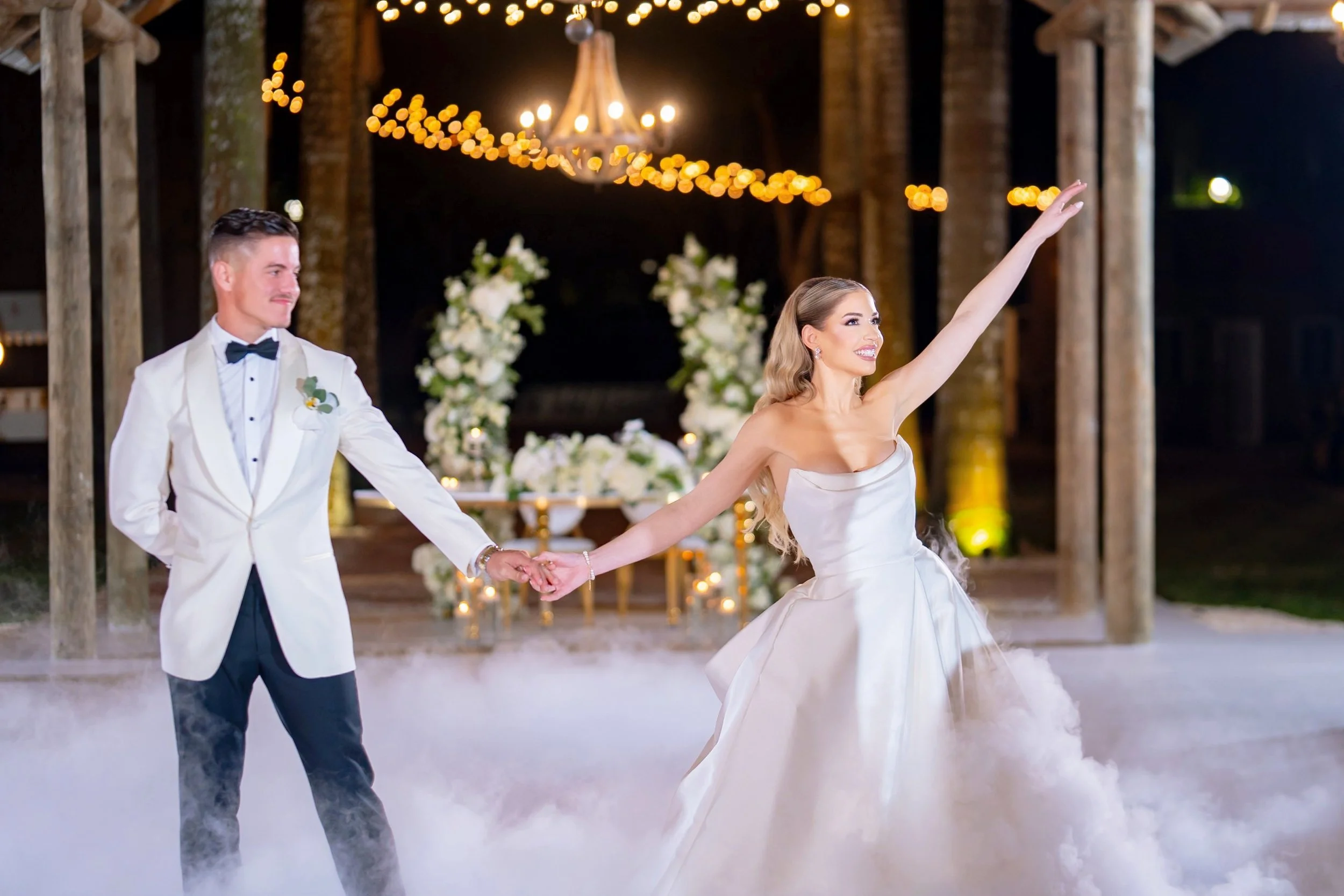 A bride and groom dancing at their wedding reception, with the bride smiling and raising her arm, and the groom holding her hand. The setting features floral arrangements, string lights, and a chandelier, with a fog effect on the dance floor.