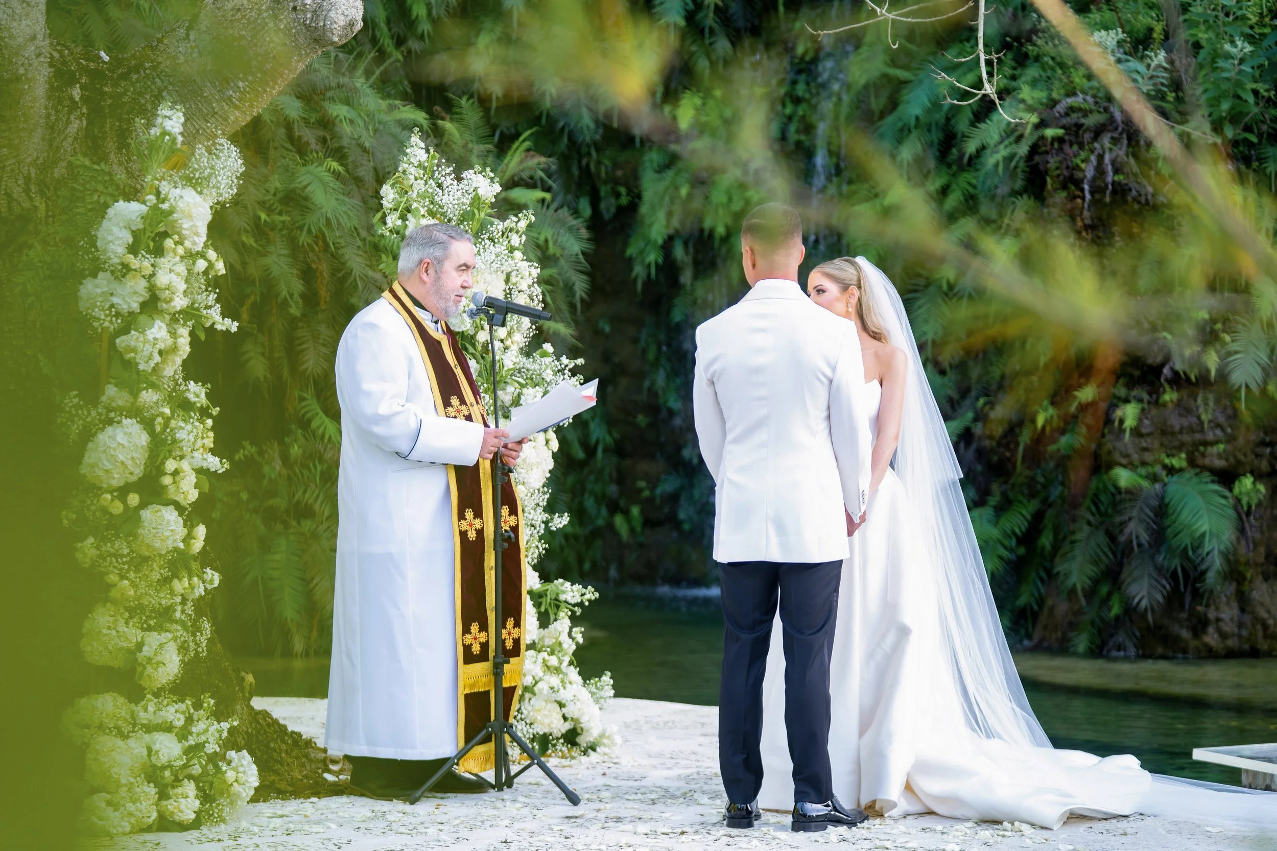 A wedding ceremony taking place outdoors, with a priest officiating, a bride in a white gown with a veil, and a groom in a white jacket, holding hands in front of a lush green background and floral decorations.