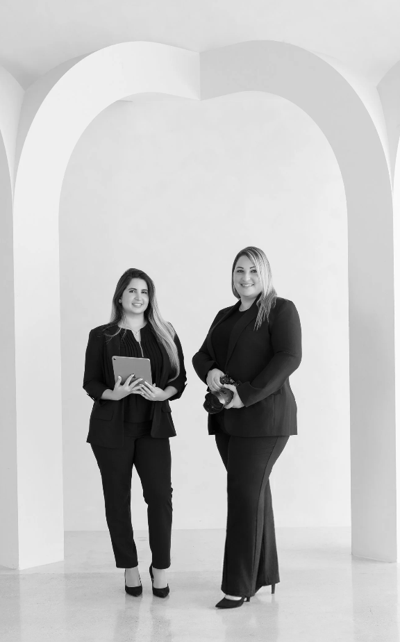 Two women in black business suits standing in front of a large white arch, smiling, one holding a tablet and the other holding a camera.