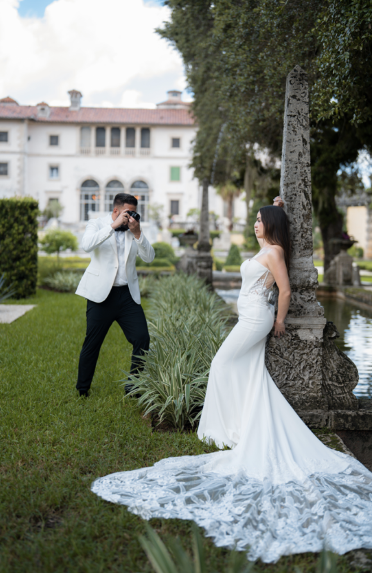 A bride in a white wedding dress posing outdoors against a stone column, while a man in a white blazer and black pants takes her photograph in a garden setting with greenery and a mansion in the background.