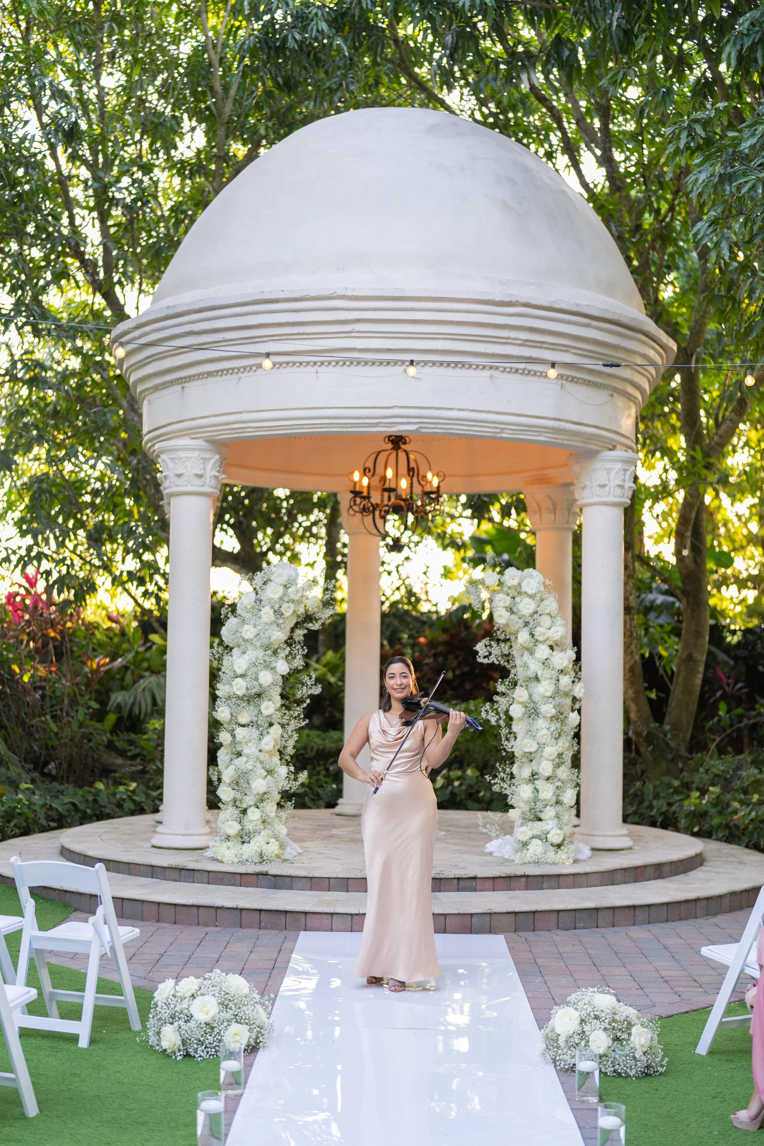 A woman in a beige gown playing the violin at an outdoor wedding ceremony under a white gazebo decorated with white flowers. chairs are arranged on either side of a white aisle runner, with flower arrangements and candles.