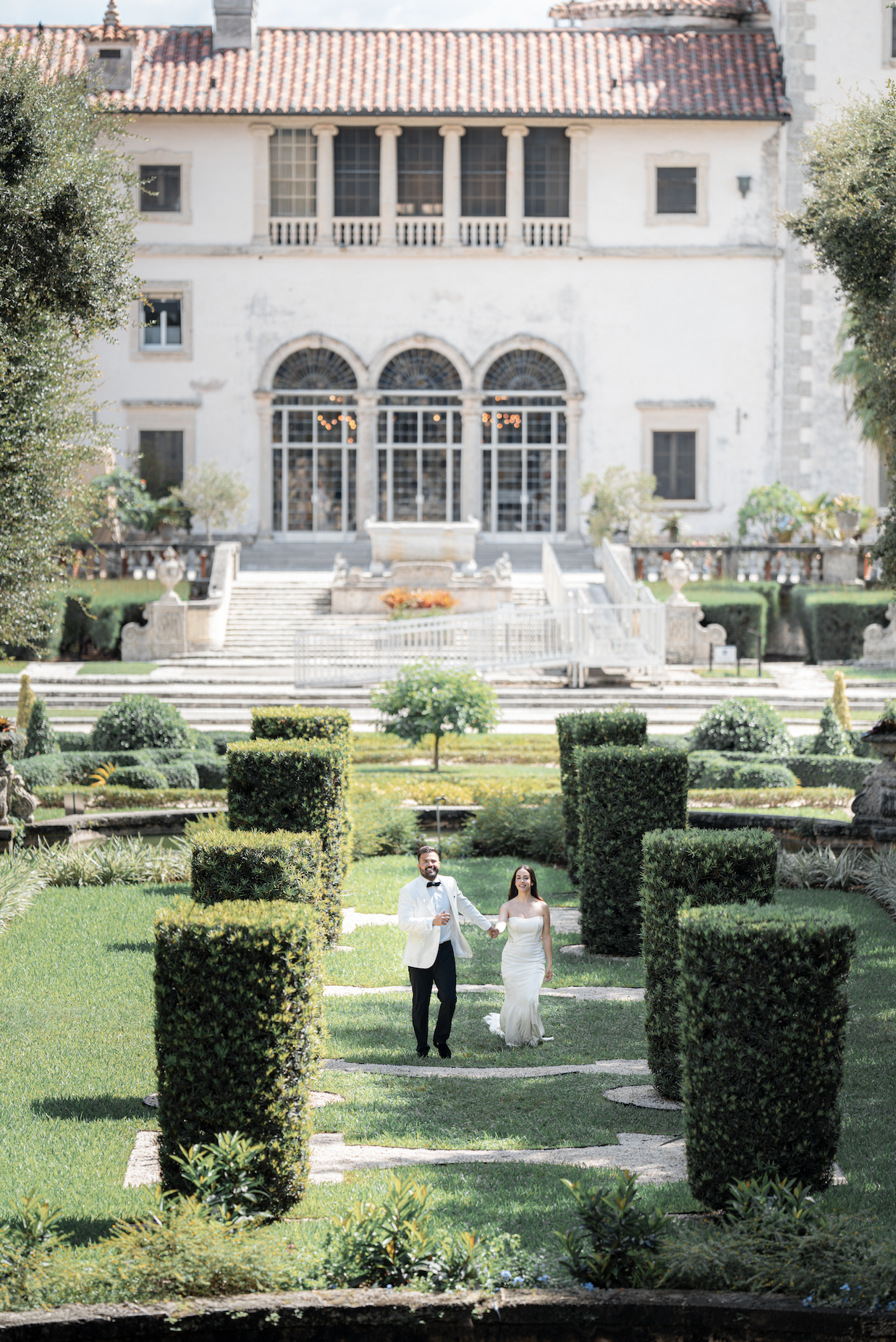 A newlywed couple holding hands and walking through a lush, manicured garden with shaped bushes and trees in front of a historic mansion.