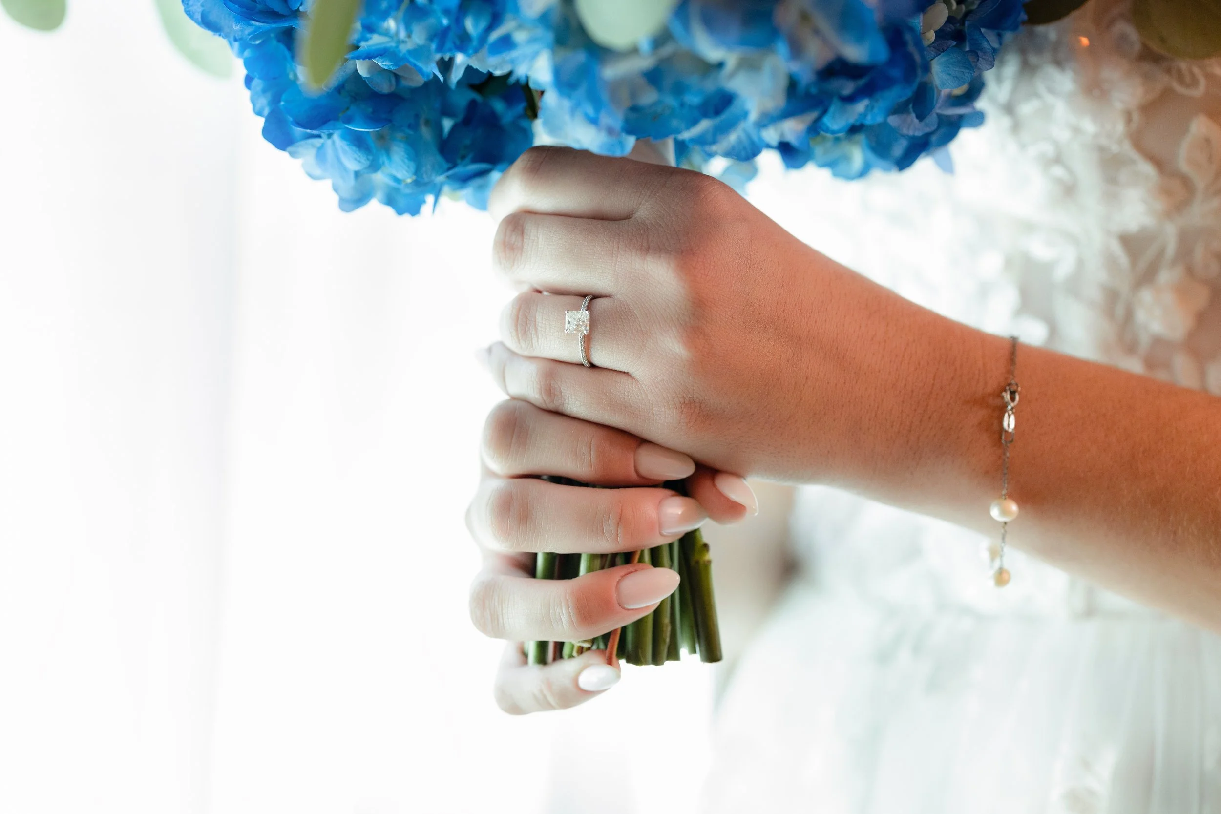 A bride holding a bouquet of blue hydrangeas, wearing a diamond engagement ring and a pearl bracelet on her wrist.