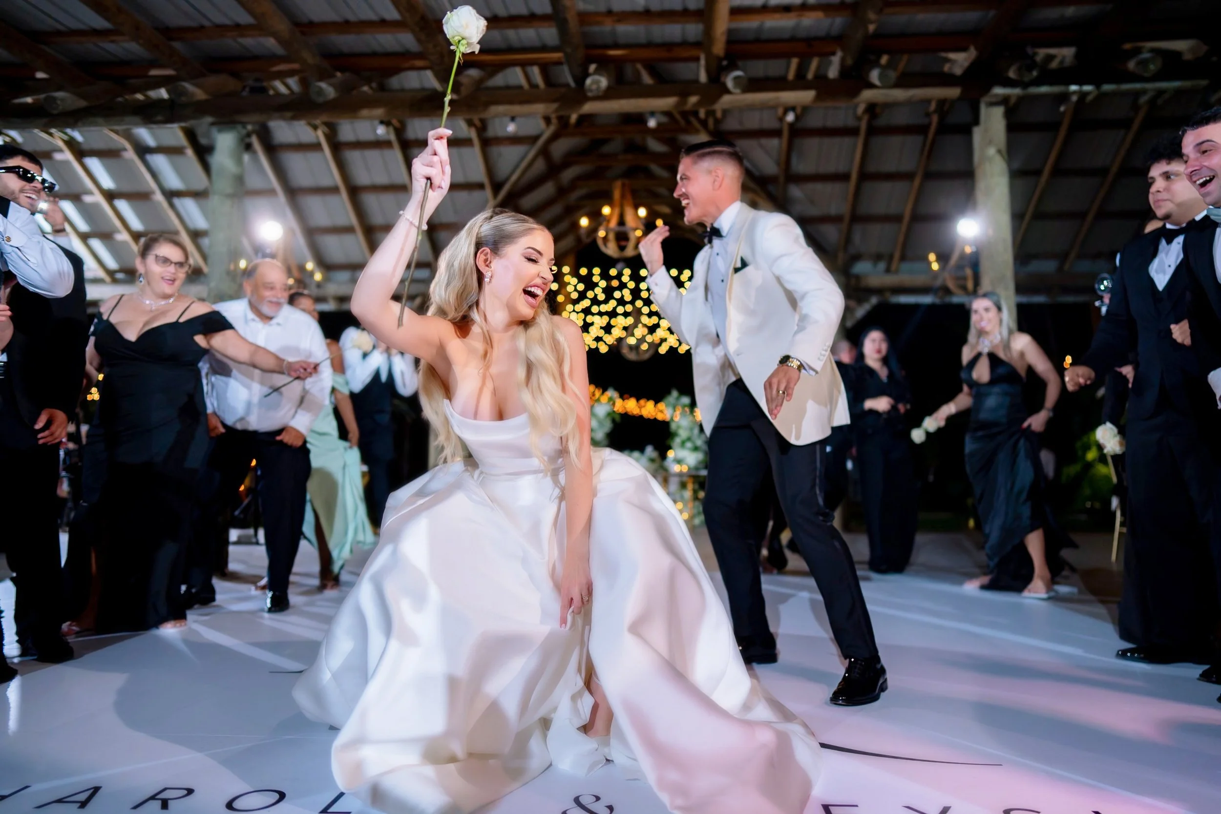 A bride in a white wedding gown happily dancing and smiling, holding a white flower on a stick, surrounded by guests in formal attire at a wedding reception in a decorated rustic venue.