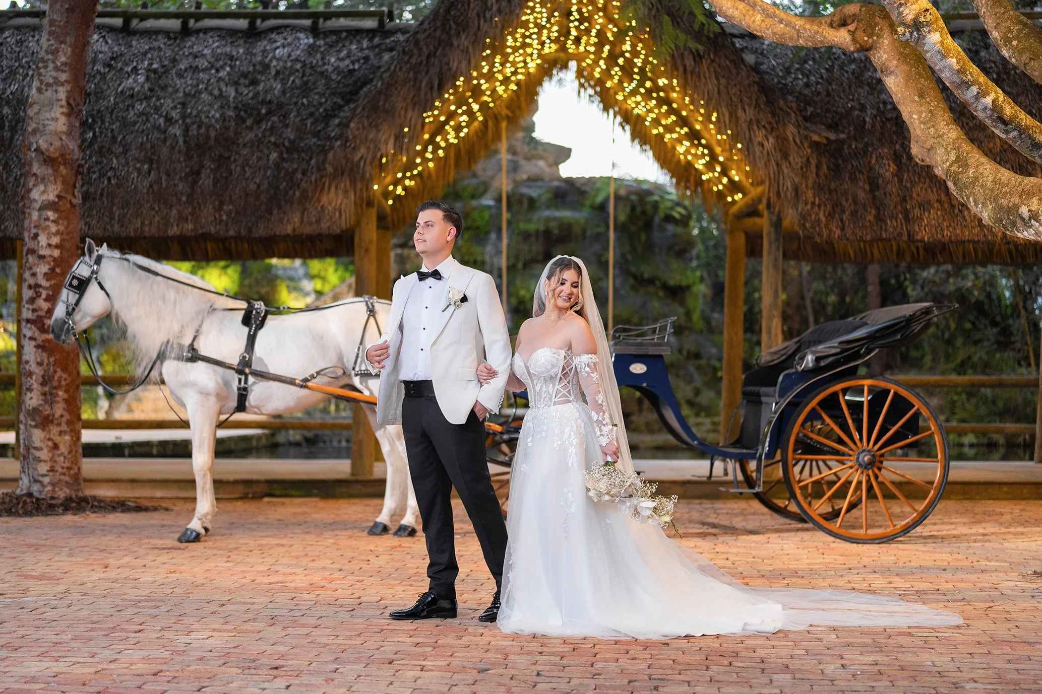 Bride and groom standing outside near a horse-drawn carriage under a thatched-roof structure decorated with string lights, with trees and natural scenery in the background.