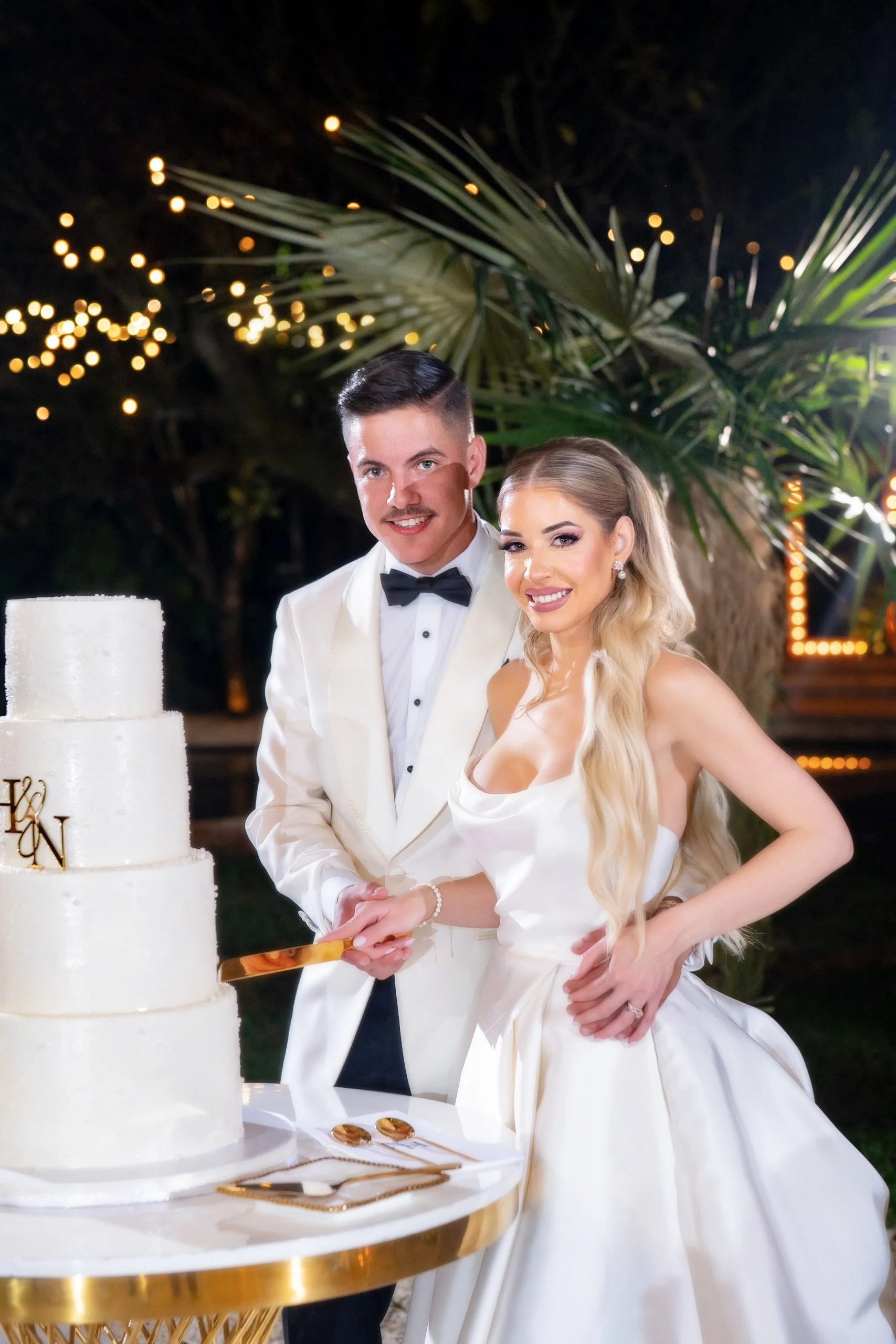 A bride and groom in wedding attire cutting their wedding cake at an outdoor celebration during the evening, with string lights and greenery in the background.