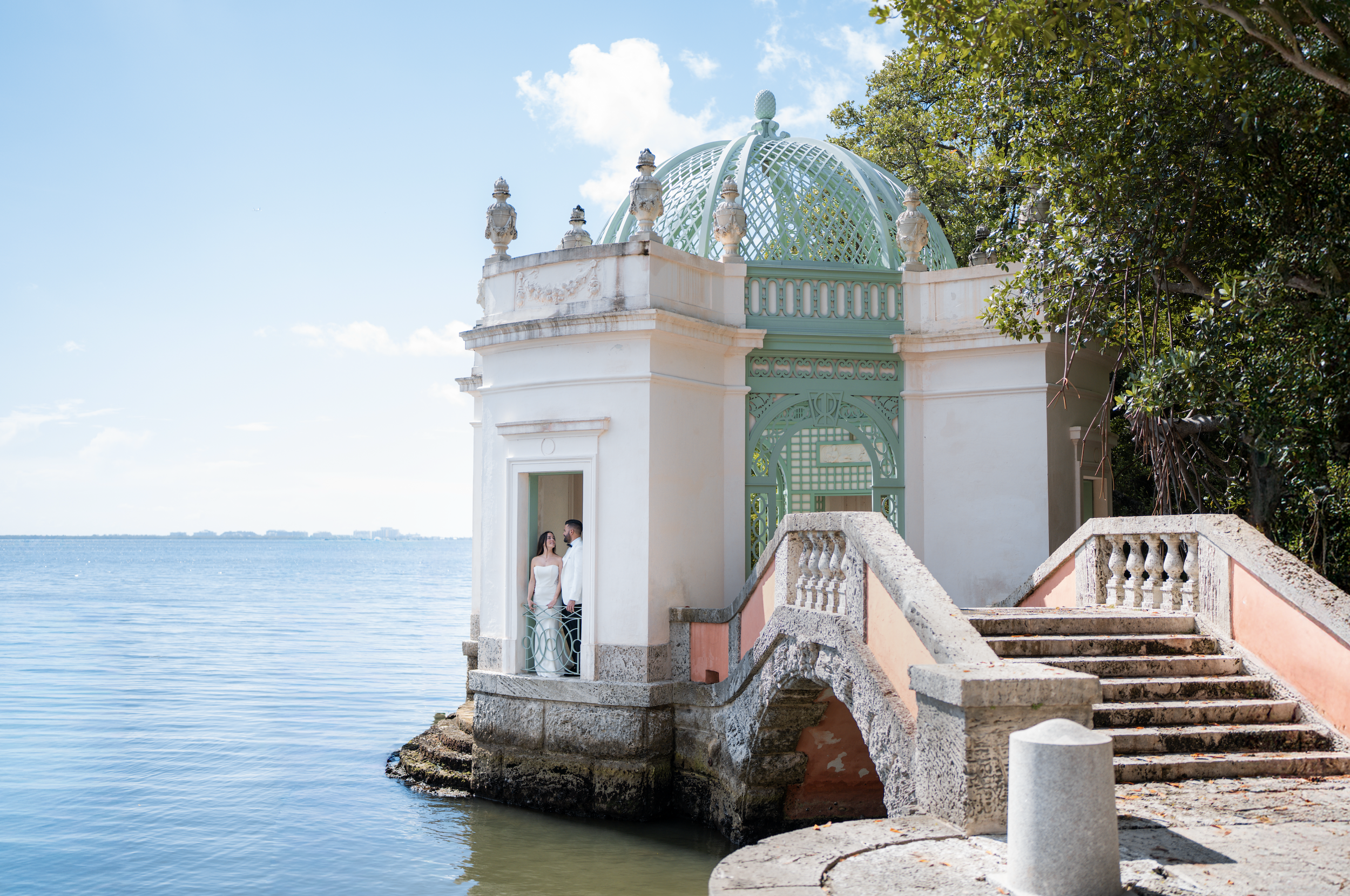 A couple standing in a small, ornate gazebo on the water's edge, with a clear sky and city skyline in the background.