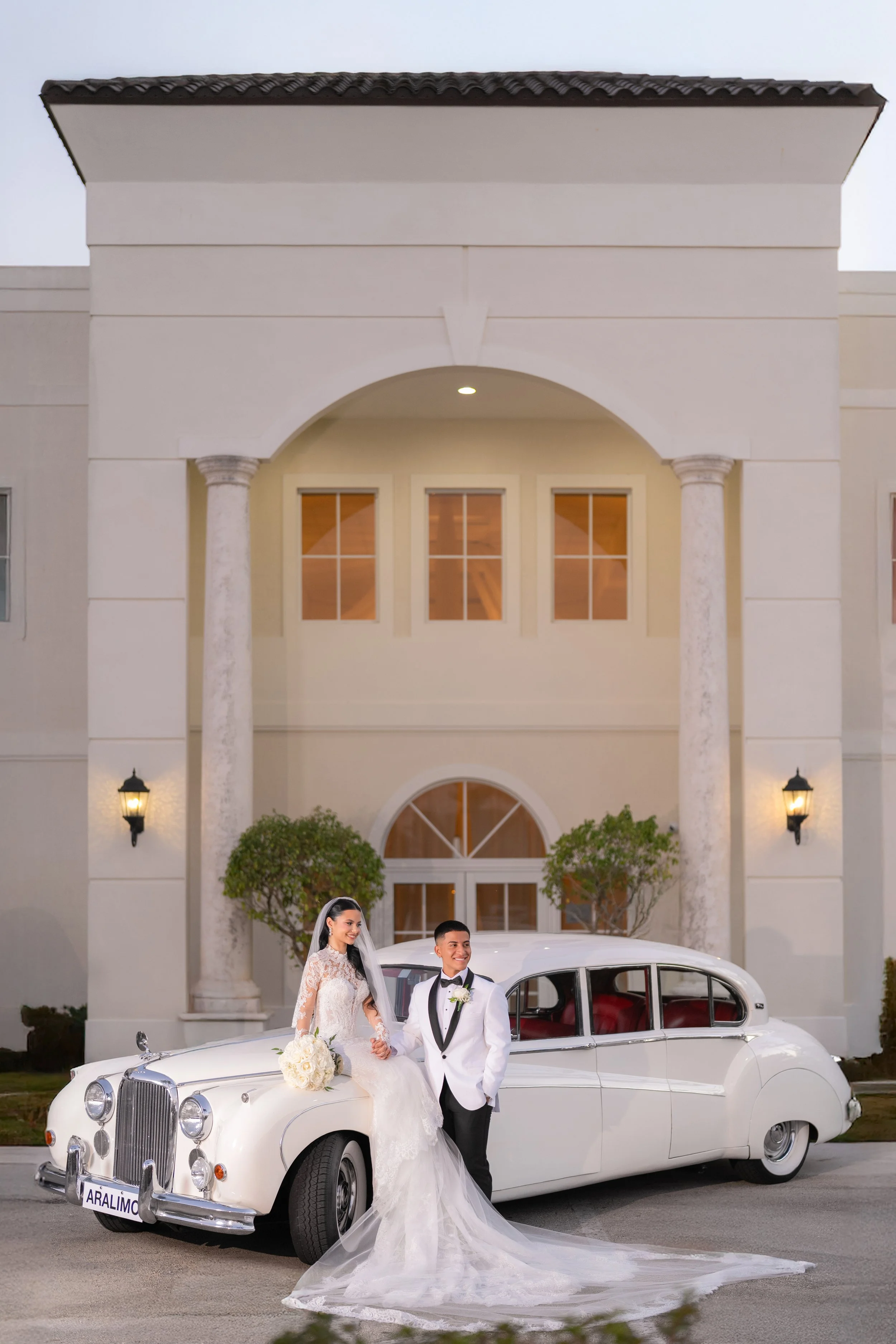 A bride and groom in wedding attire standing beside a white vintage car in front of a grand building with columns and lit wall sconces.