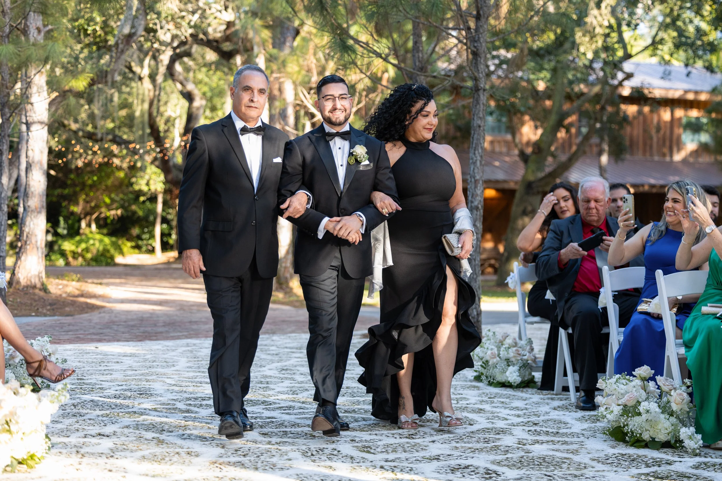 A wedding procession walking down an outdoor aisle surrounded by trees and guests seated on white chairs, with some guests taking photos.
