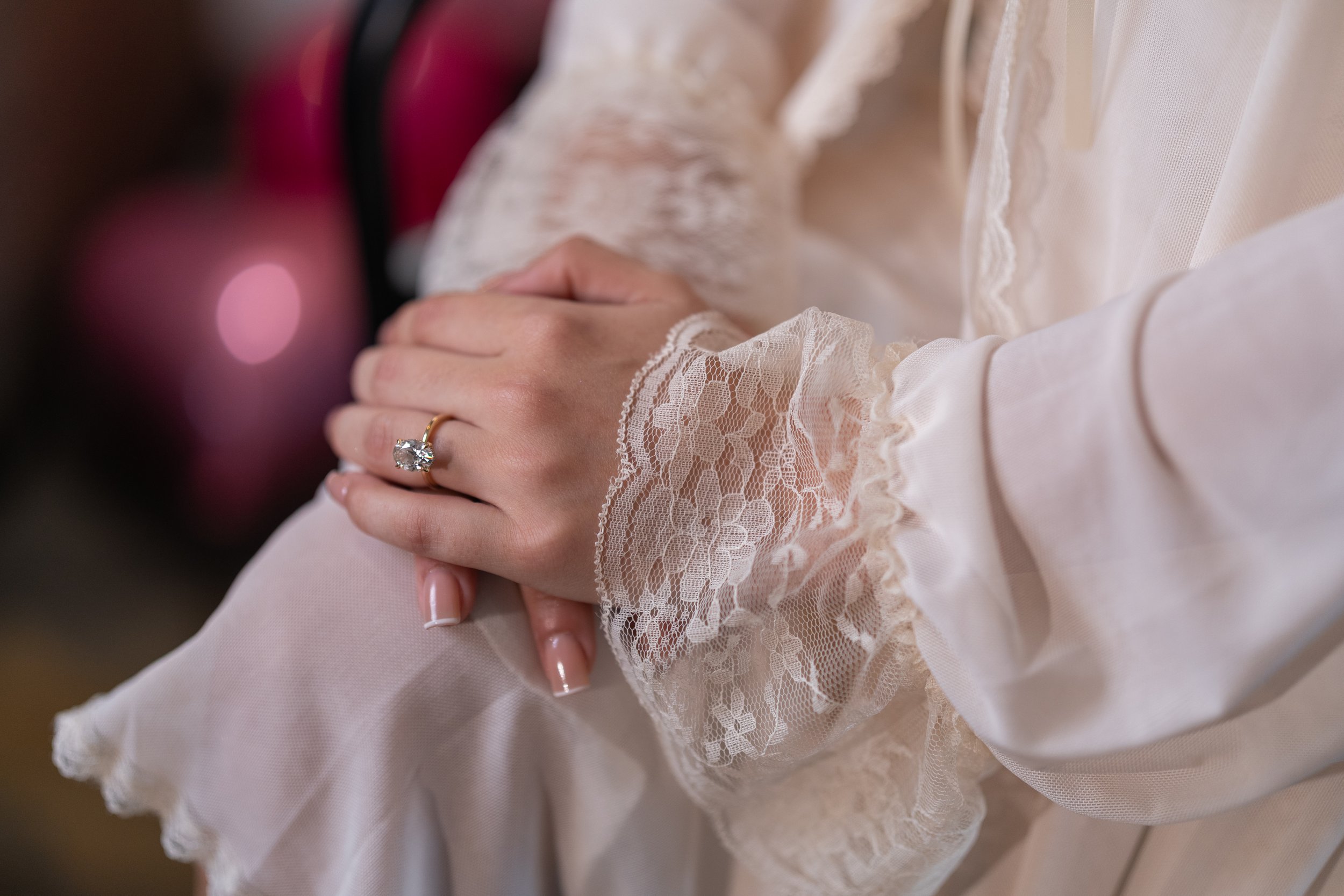Close-up of a woman's hands resting on her lap, wearing a lace-cuffed, cream-colored blouse and a large diamond engagement ring.