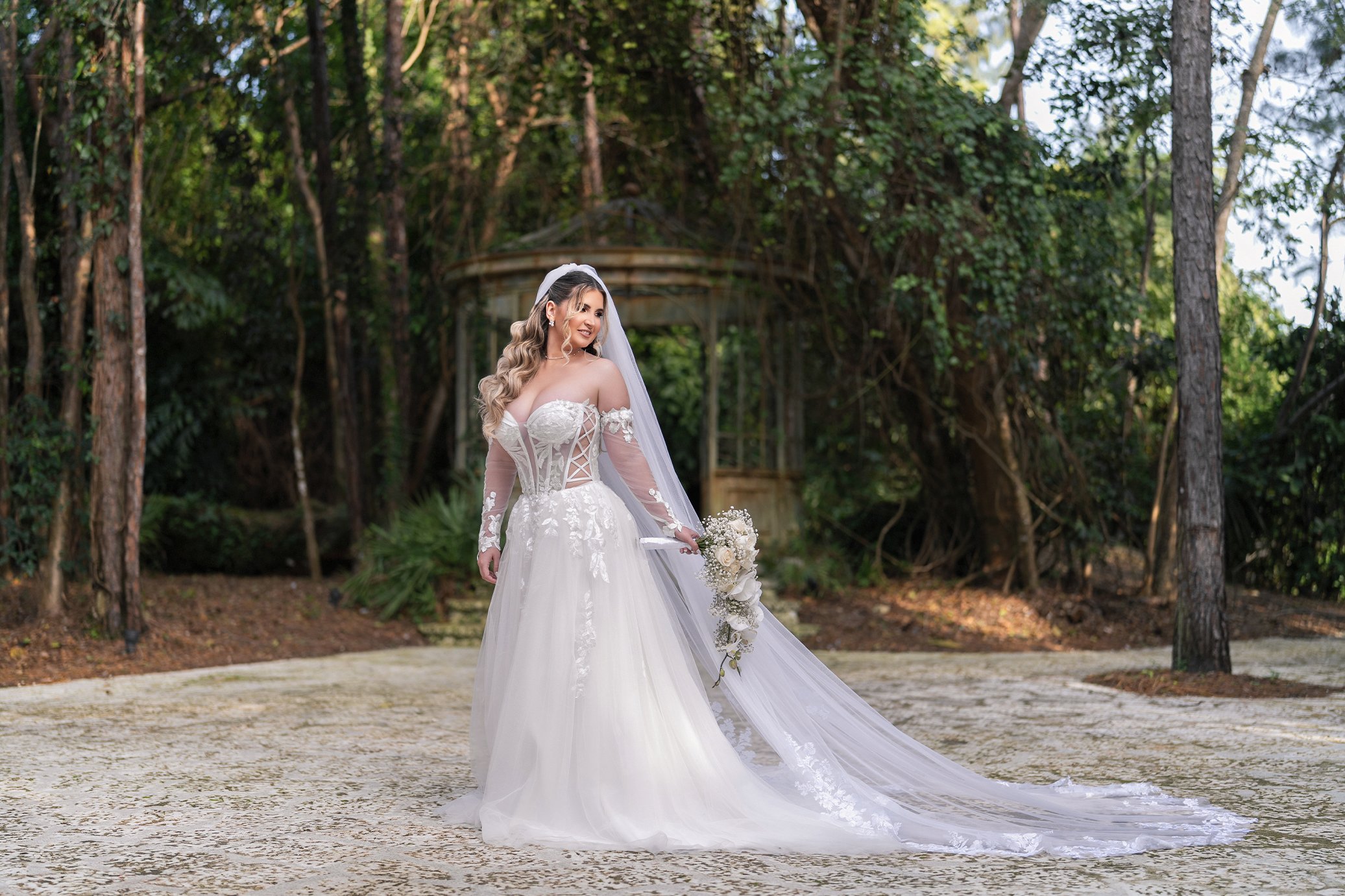 Bride in a white wedding gown holding a bouquet of flowers standing outdoors in front of a wooded area with a gazebo in the background.
