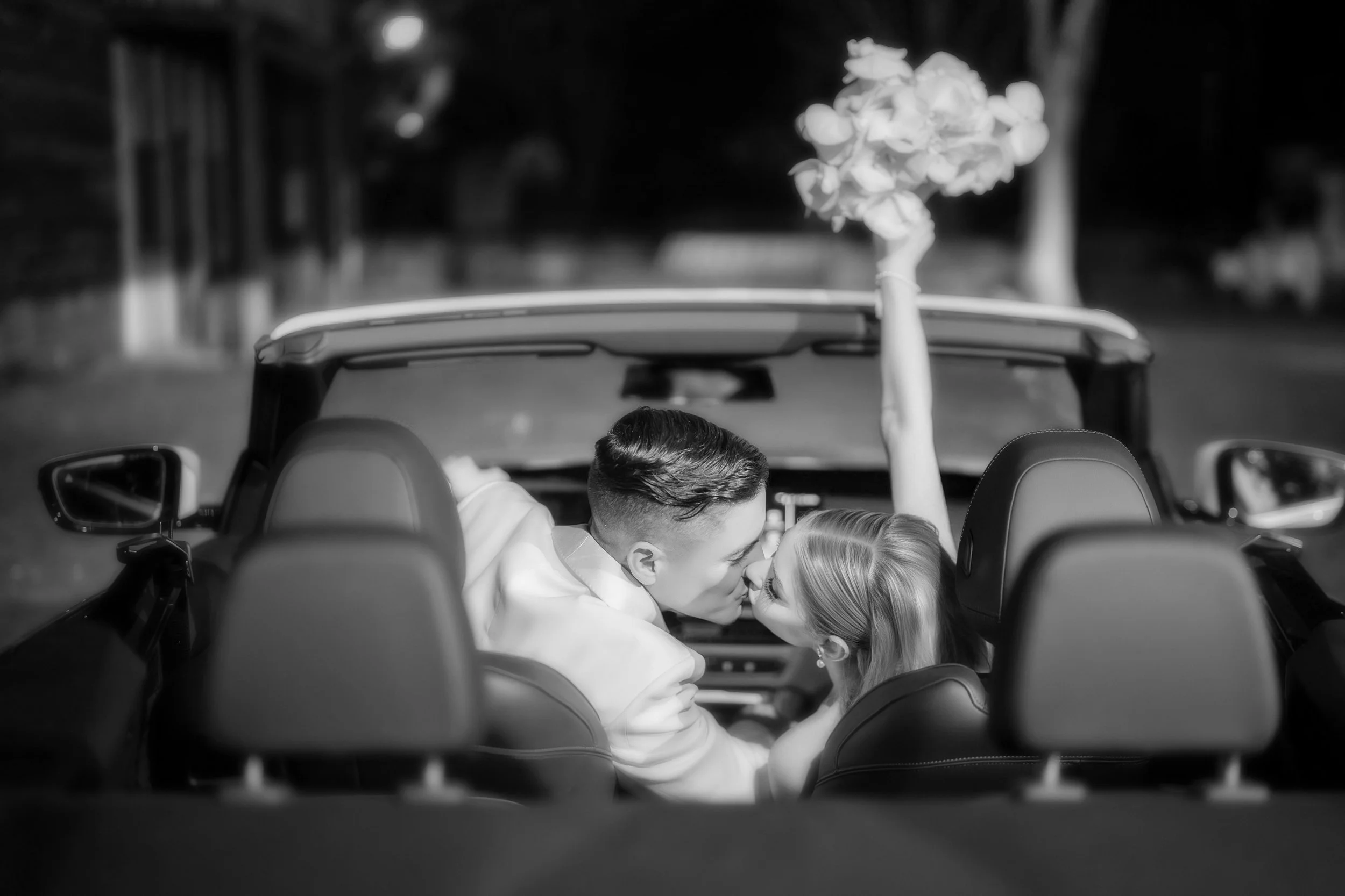 A black-and-white photo of a couple kissing in a convertible car at night, with a bouquet of flowers attached to the car's window.