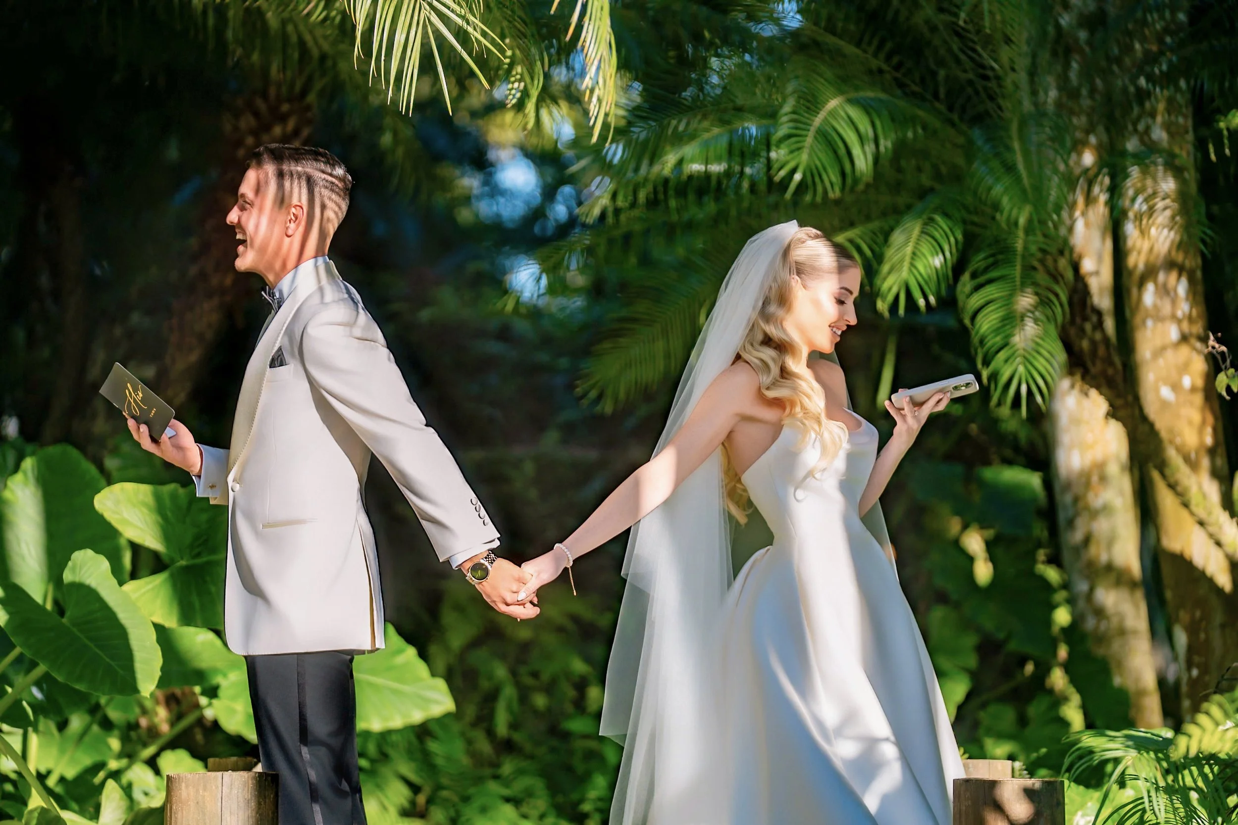 A bride and groom are standing outdoors in a lush green garden, holding hands with their backs touching. Both are smiling and appear joyful. The groom is wearing a white tuxedo and holding a smartphone, while the bride is in a white wedding gown with