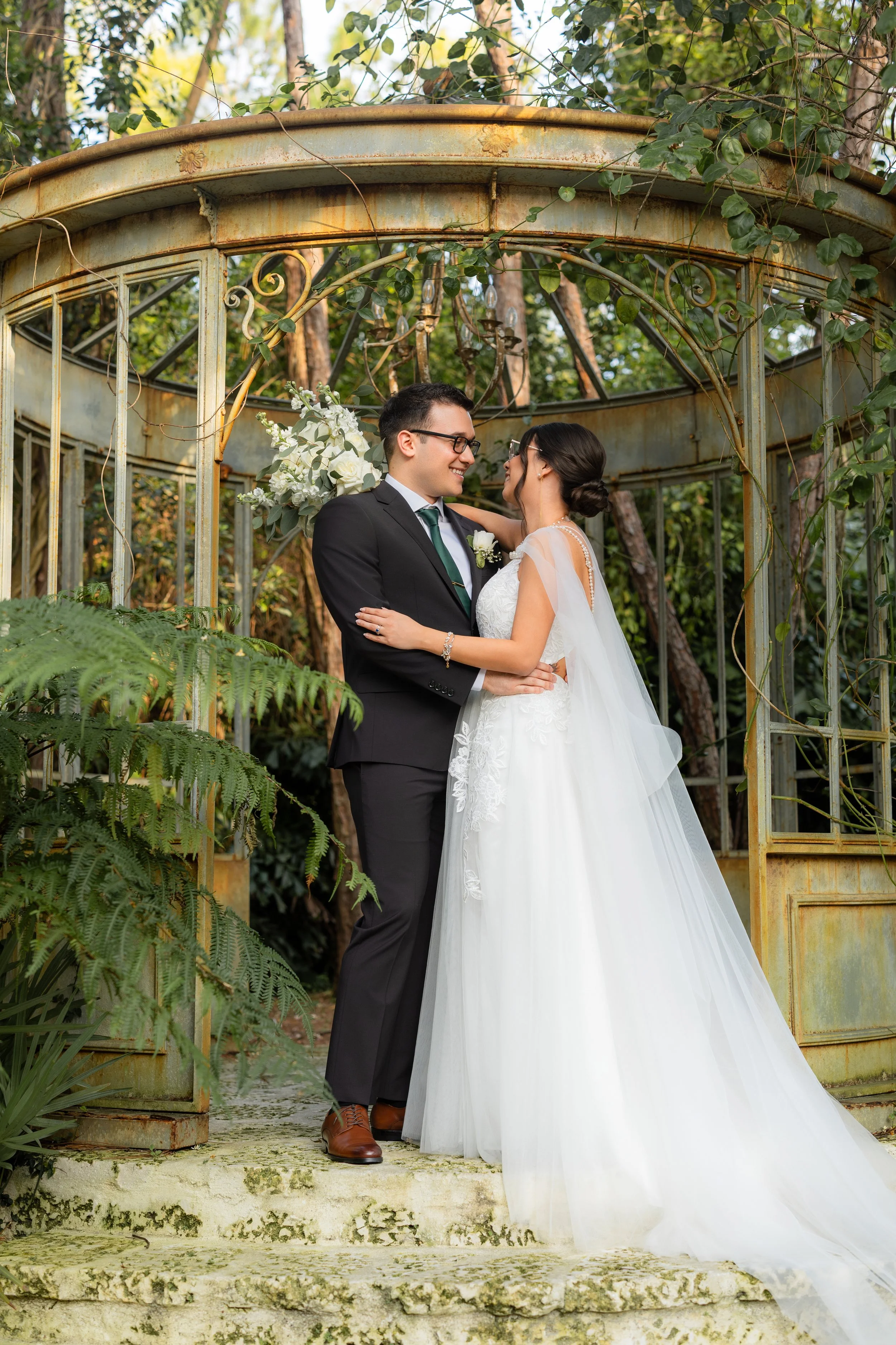 A bride and groom share a kiss and smile at each other, standing on moss-covered steps in front of a rusted, vintage greenhouse structure surrounded by lush greenery.