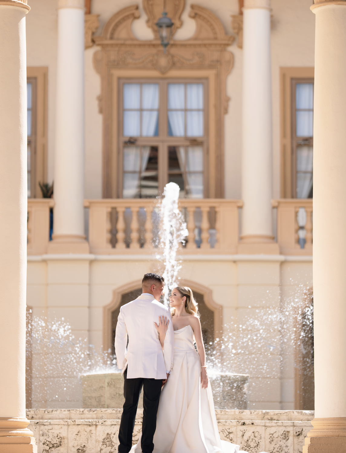 A bride and groom stand close together in front of a fountain on their wedding day, with the bride smiling at the groom. The fountain is in front of a grand building with large windows, columns, and ornate woodwork details.