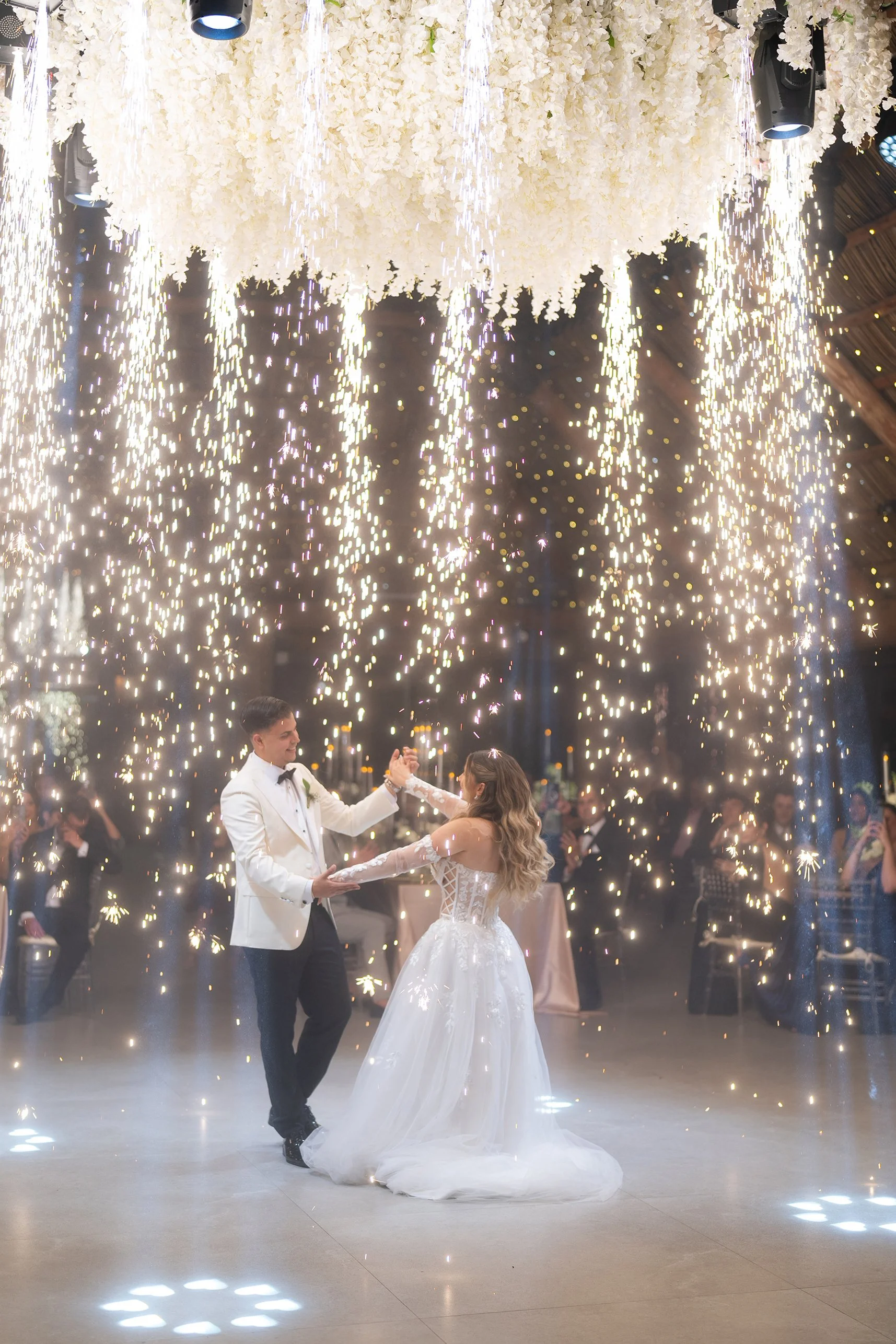 A bride and groom dancing at their wedding reception under a shower of sparks and a floral chandelier ceiling.